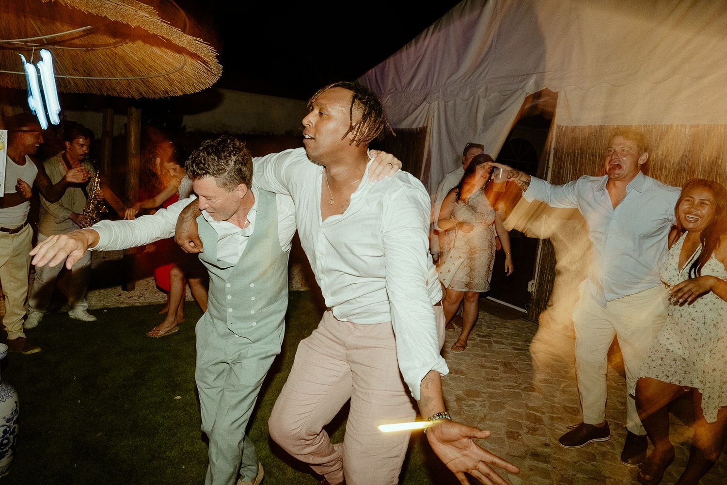 Two men dancing joyfully at an outdoor wedding reception party at night with guests celebrating.