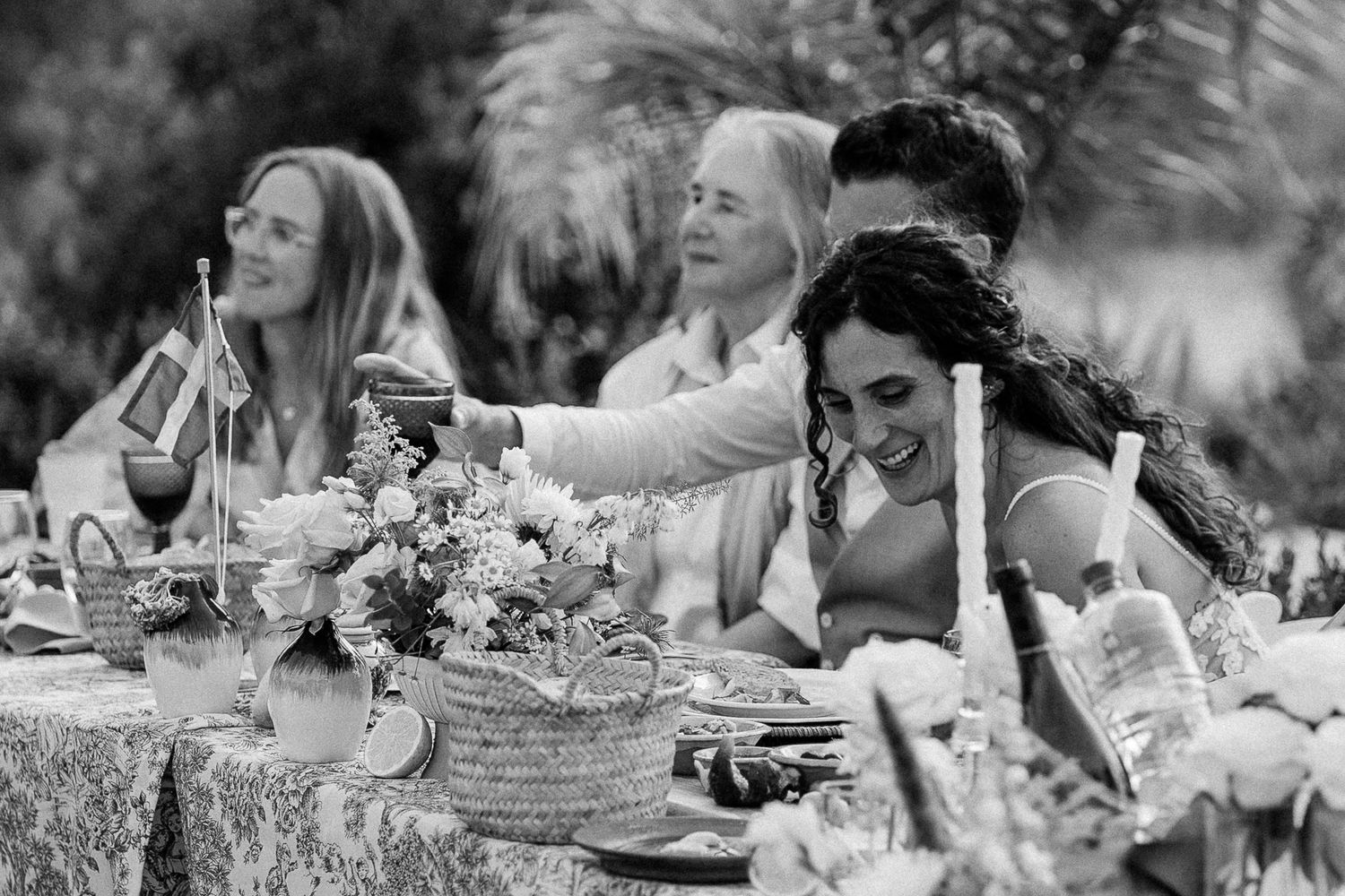 Black and white photo of guests laughing and toasting at an outdoor garden wedding reception dinner table.