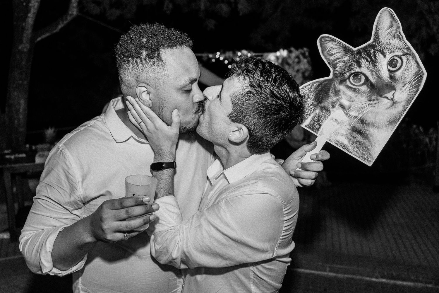 Two men sharing a kiss at a celebration, one holding a cat face prop, in black and white photography.
