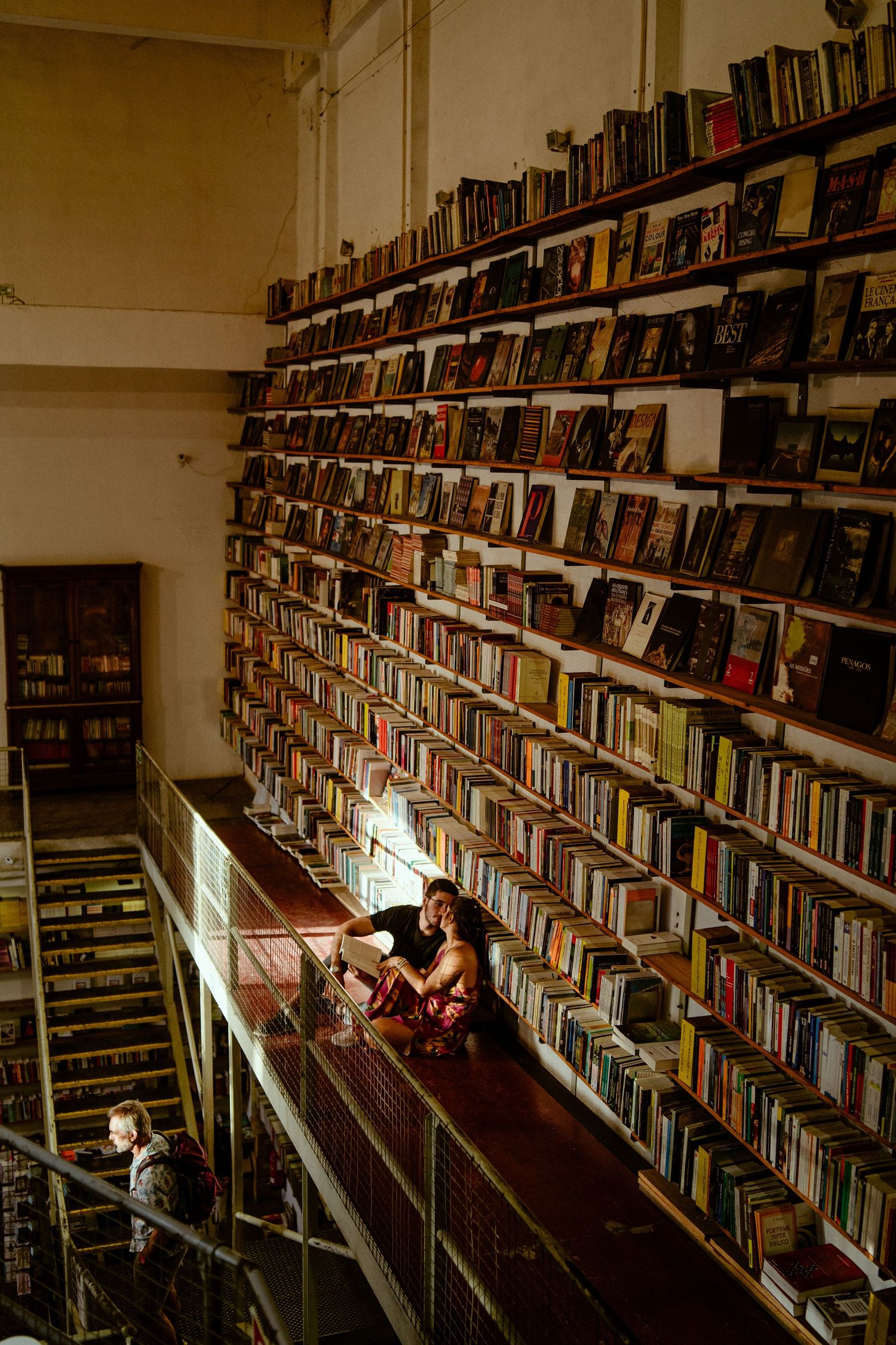 Person reads while sitting between long rows of library bookshelves filled with books in a dimly lit space.