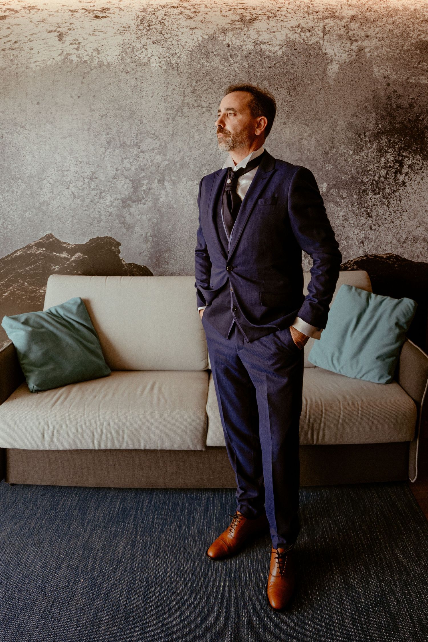 A figure in a navy suit and brown shoes sits contemplatively on a gray couch against a textured wall.