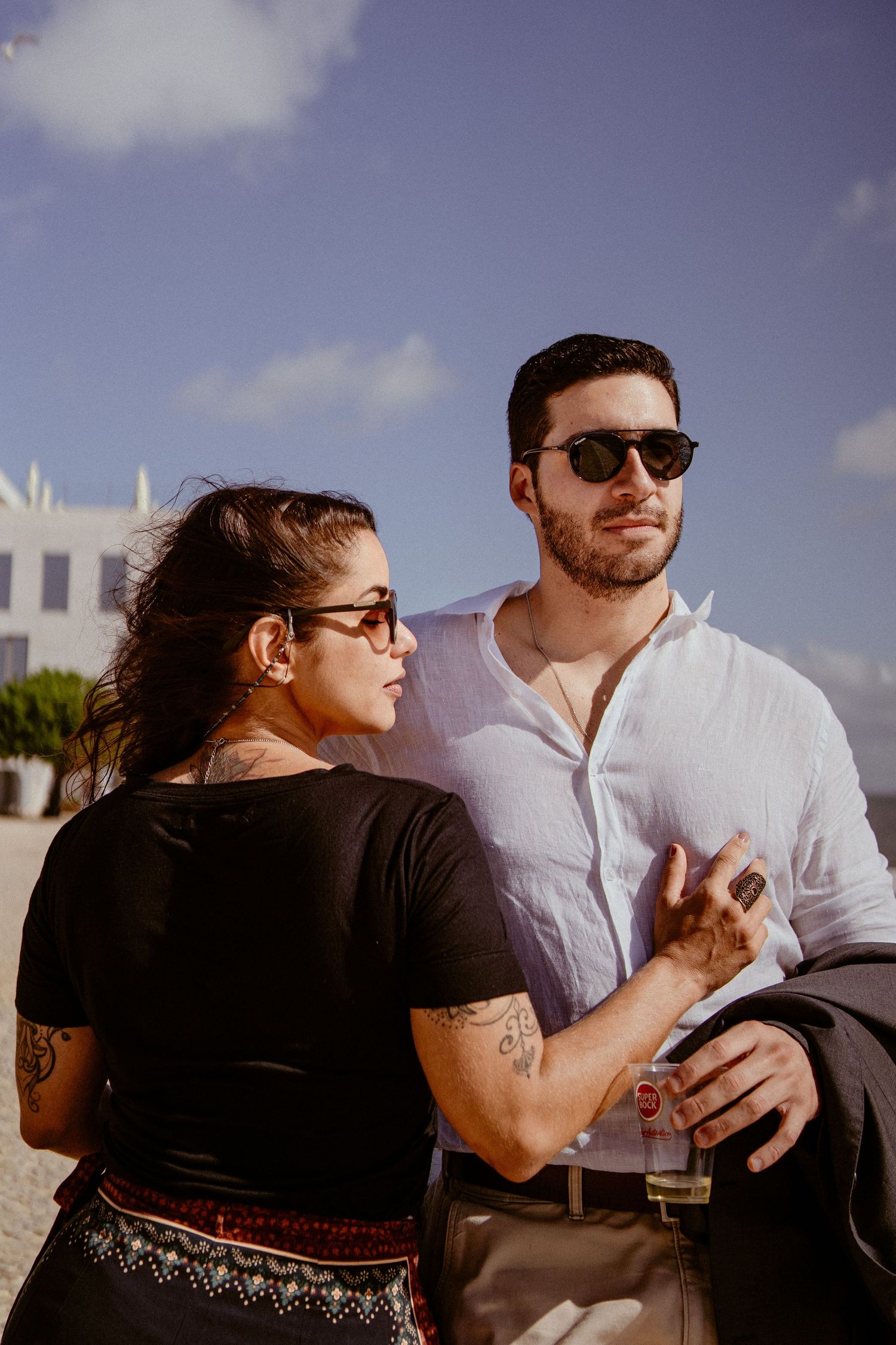 A couple sits together outdoors sharing drinks while enjoying a sunny day with blue sky and clouds in the background.