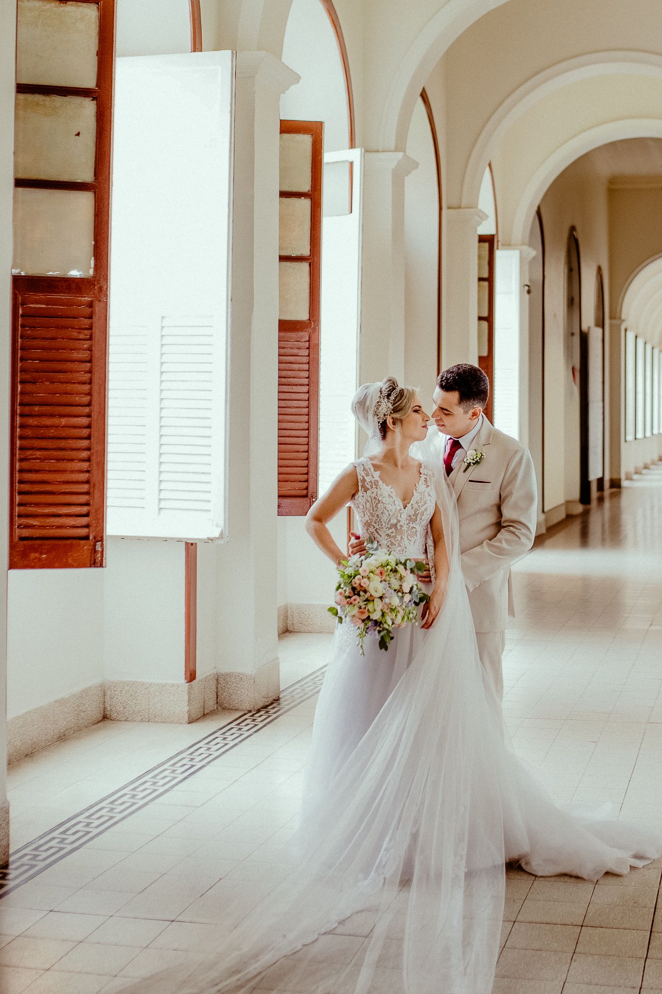 A couple in wedding attire walks through an elegant white hallway with arched columns and wooden shutters.