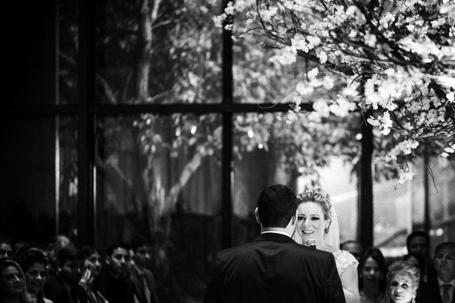 A black and white wedding ceremony photo in front of a large window with natural light streaming in.