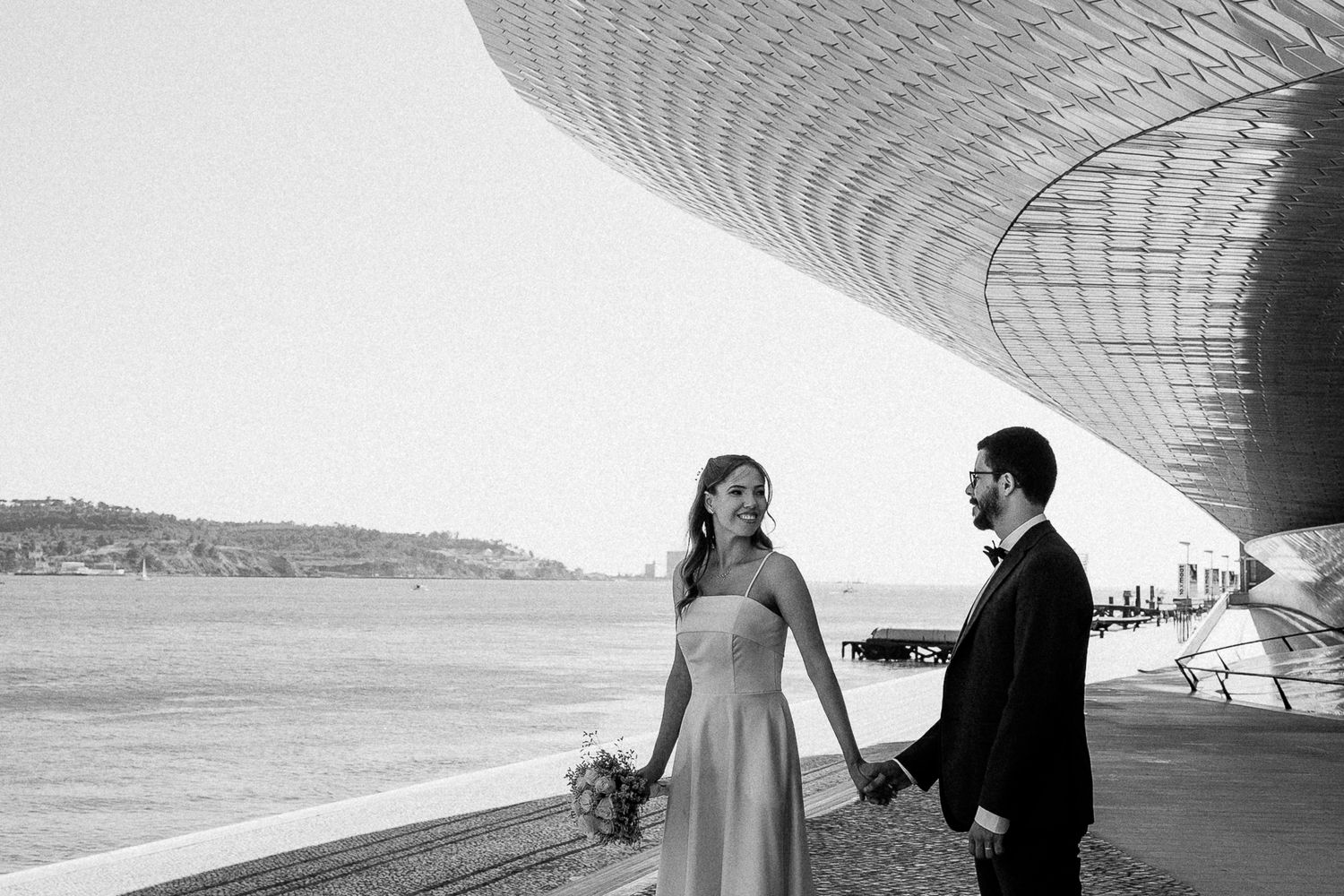 Black and white photo of a bride and groom holding hands by a modern waterfront building on their wedding day.