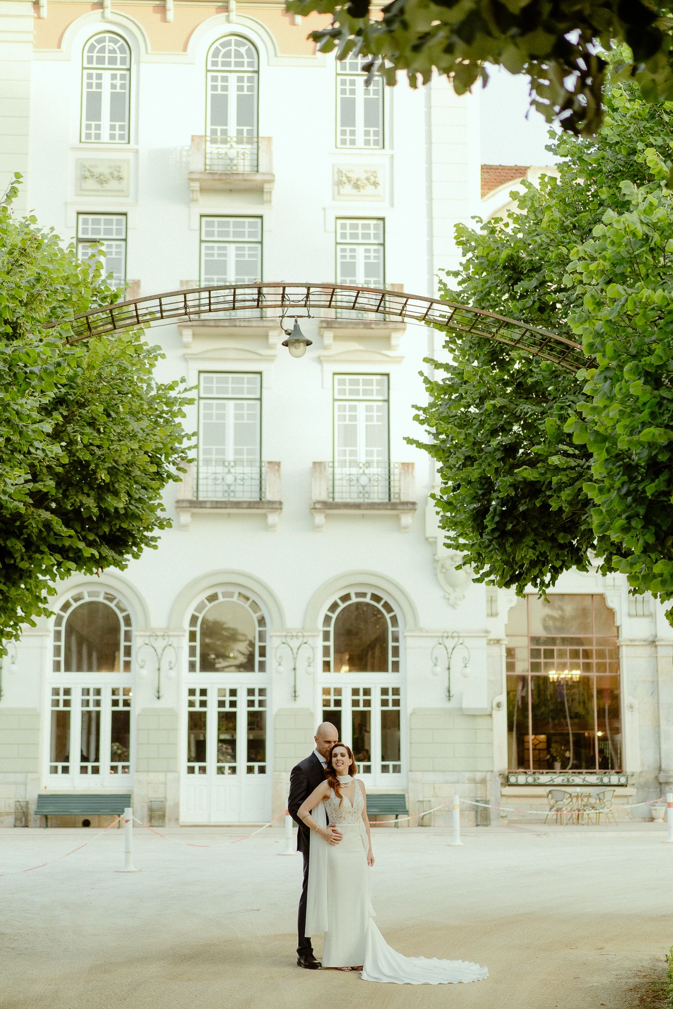 Bride and groom embracing in front of elegant white European building with arched windows and lush green trees.
