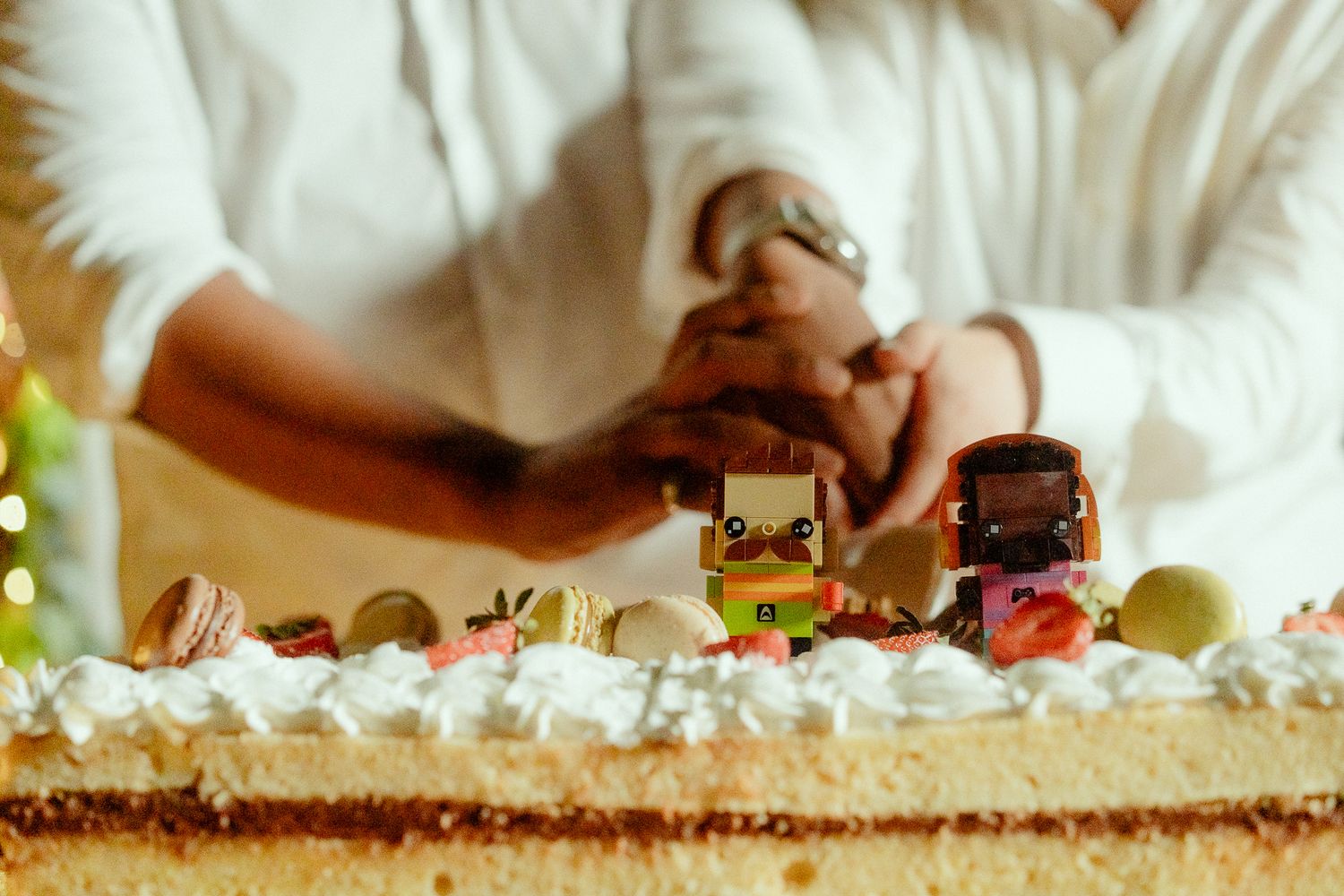 Two people cutting a decorated cake topped with LEGO figures, macarons, and whipped cream at a celebration.