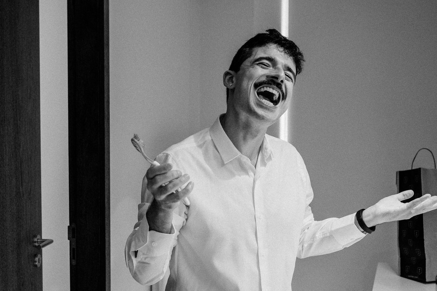 Black and white photo of a laughing man in white shirt holding a toothbrush with arms outstretched.