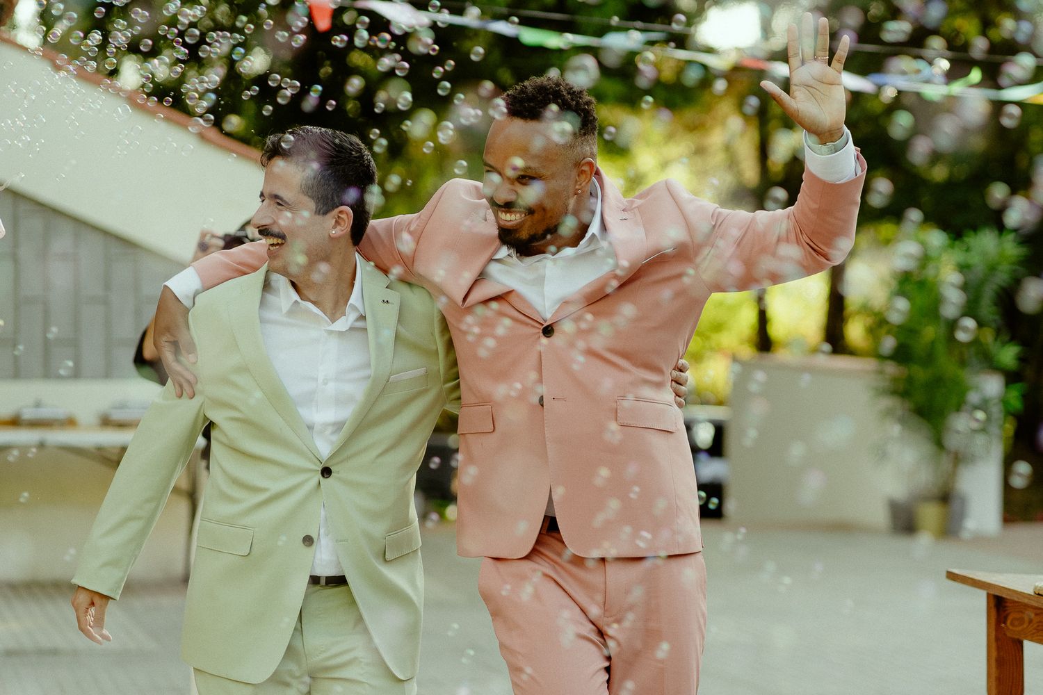 Two grooms in pastel suits celebrate joyfully with bubbles at their outdoor wedding ceremony.