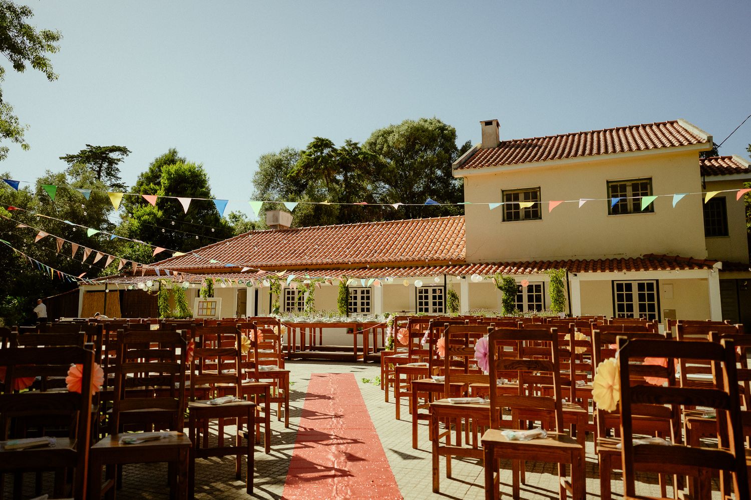 Outdoor wedding ceremony setup with wooden chairs, colorful bunting, and a rustic white venue with red tile roof.