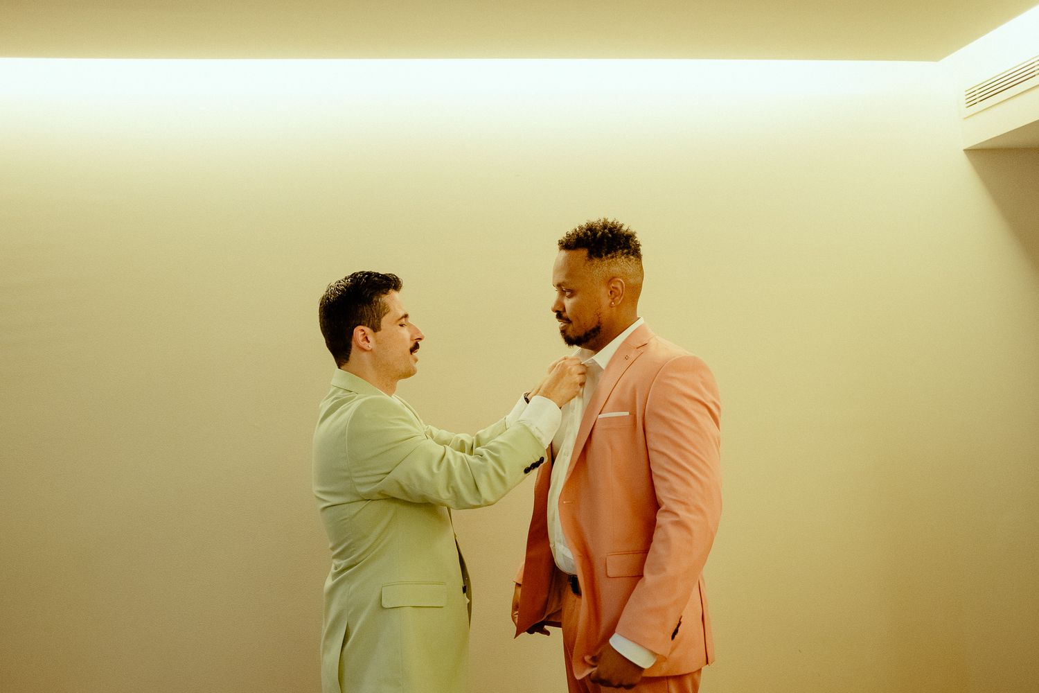 Two men in pastel suits adjusting neckwear, one in sage green helping the other in a pink suit.