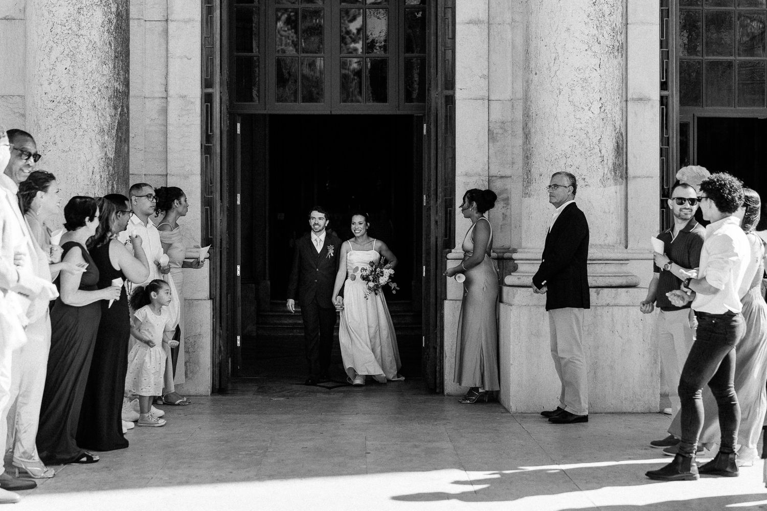 Bride and groom exit a grand building as wedding guests line up outside in this classic black and white photo.