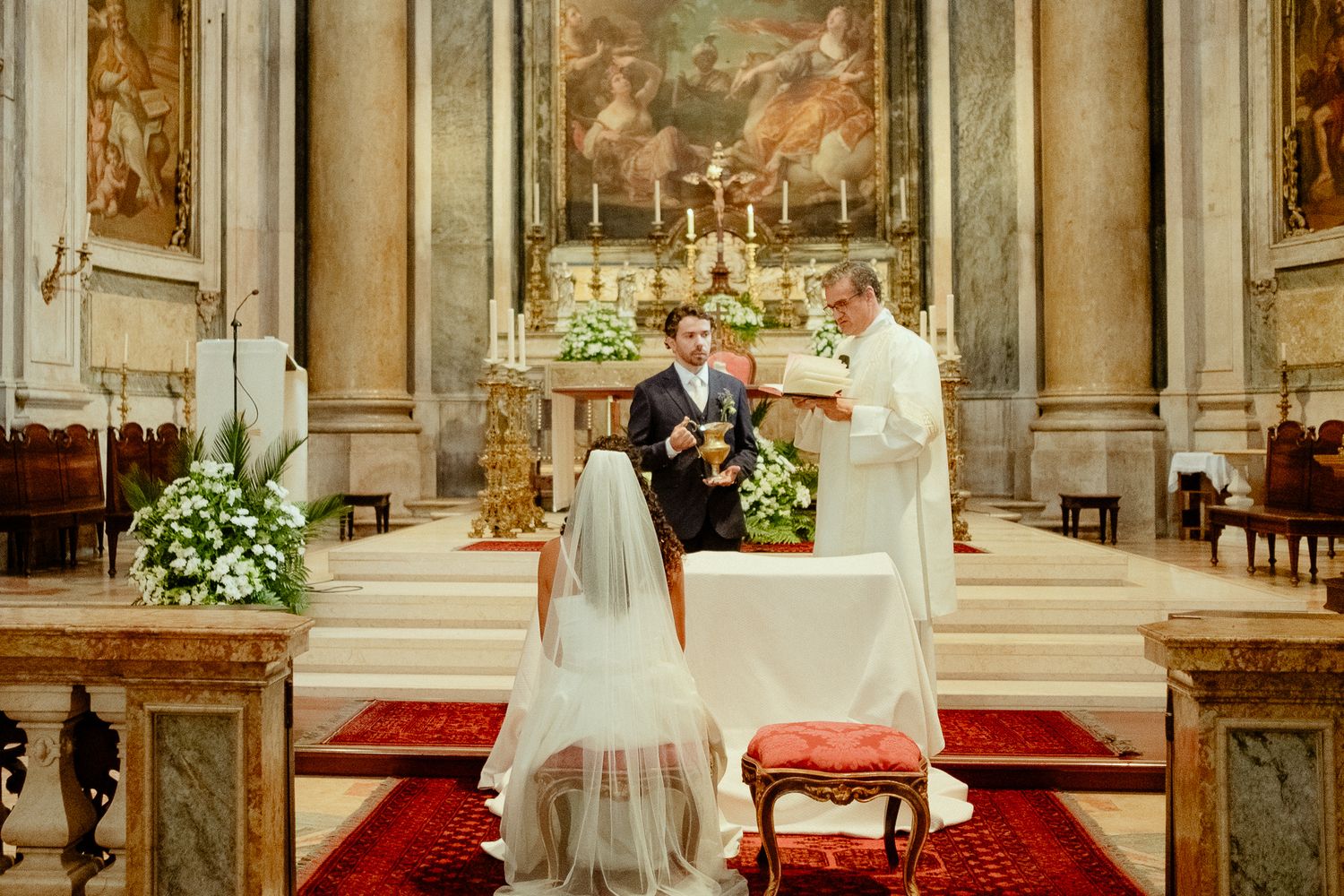 Bride and groom exchange vows at a Catholic church altar adorned with floral arrangements during a traditional wedding ceremony.