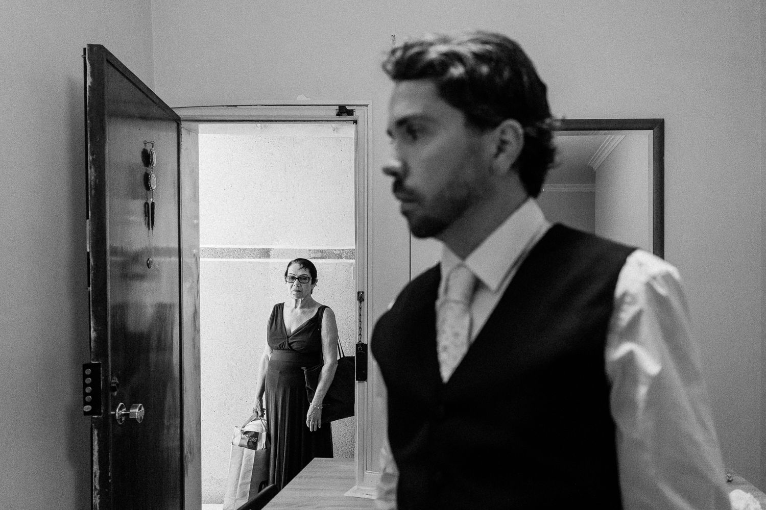 Black and white photo of a man in vest and woman in doorway during a wedding preparation moment.