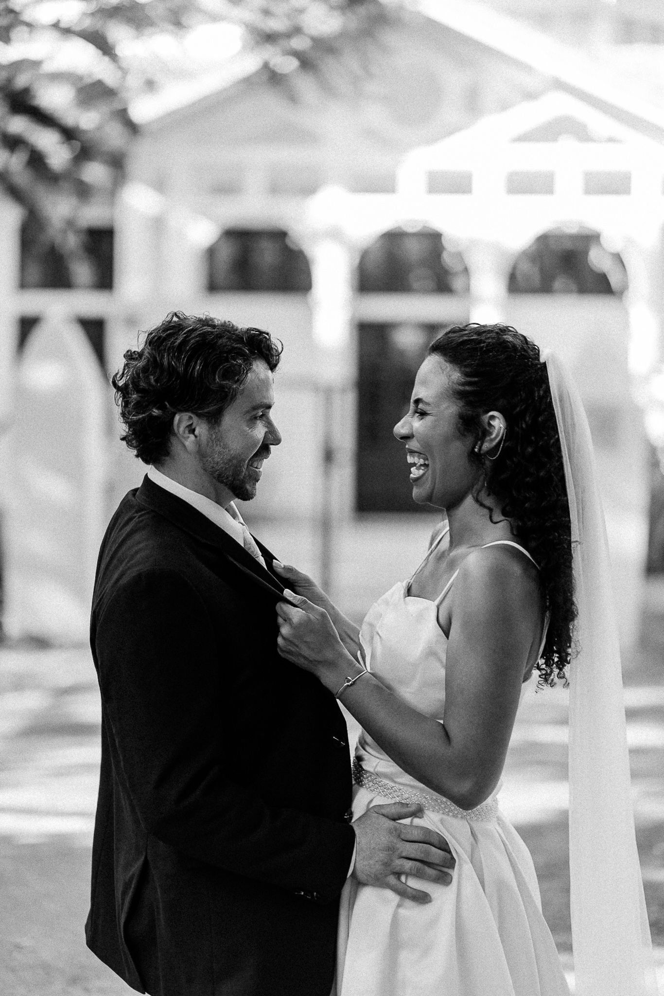 Black and white wedding photo of smiling bride in veil and groom in suit sharing a tender moment outdoors.
