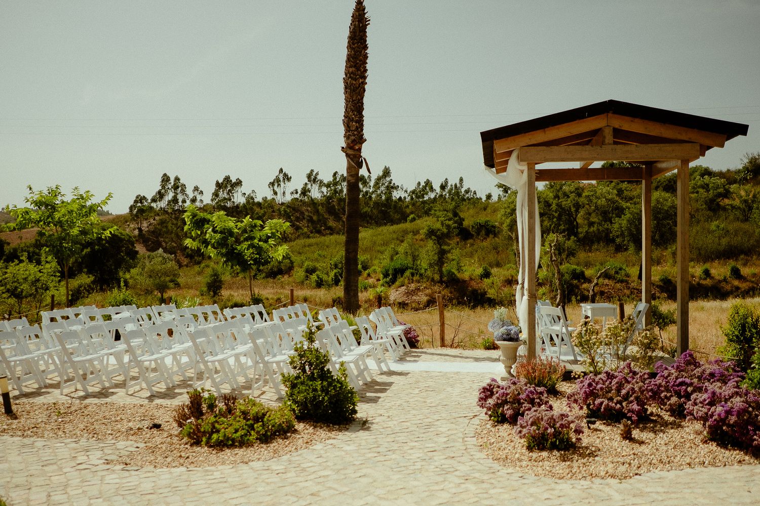 Outdoor wedding ceremony setup with white chairs, floral arbor, and lush greenery backdrop on a sunny day.