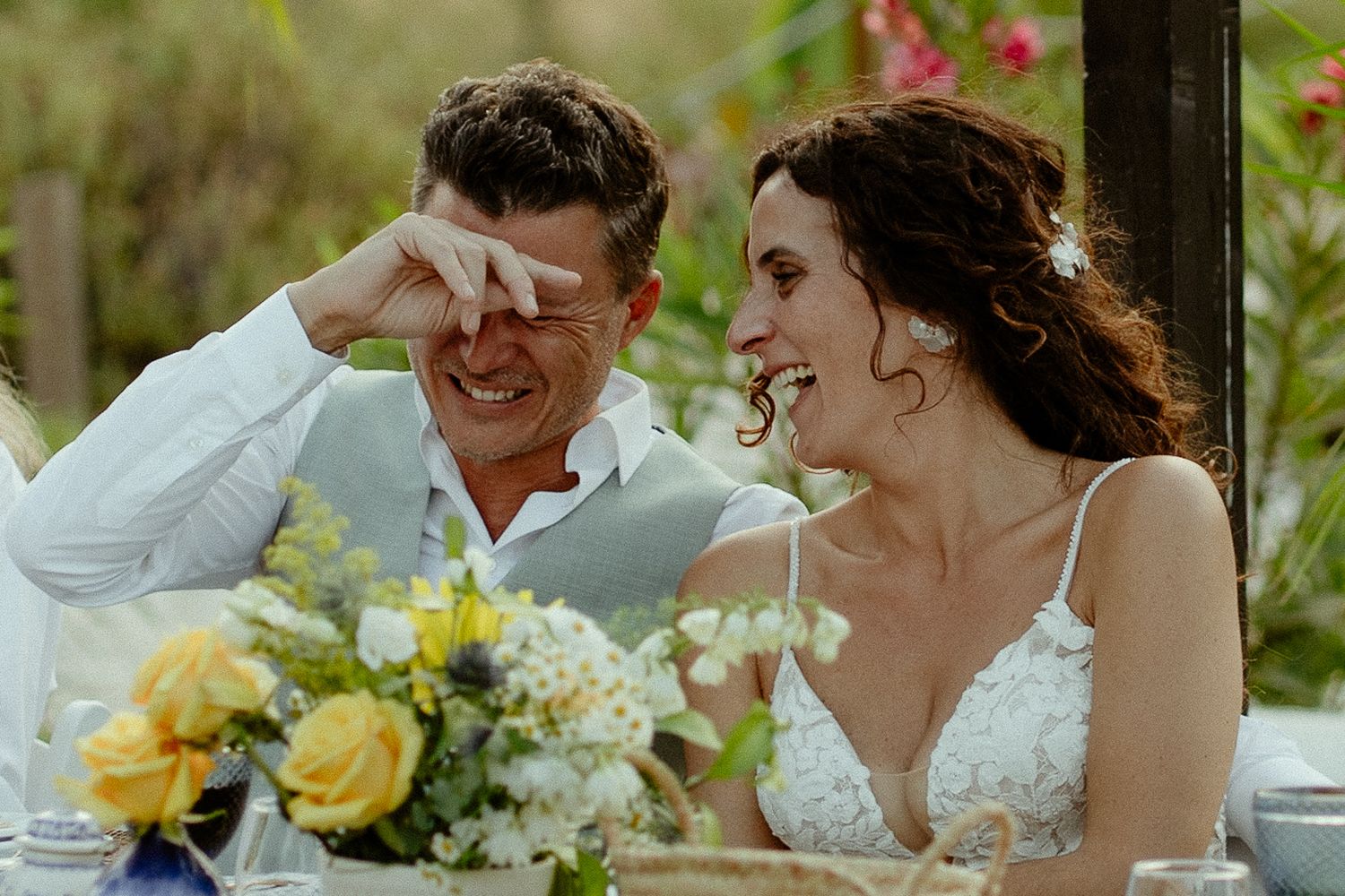 Bride and groom laughing joyfully at wedding reception table with yellow and white floral centerpiece.