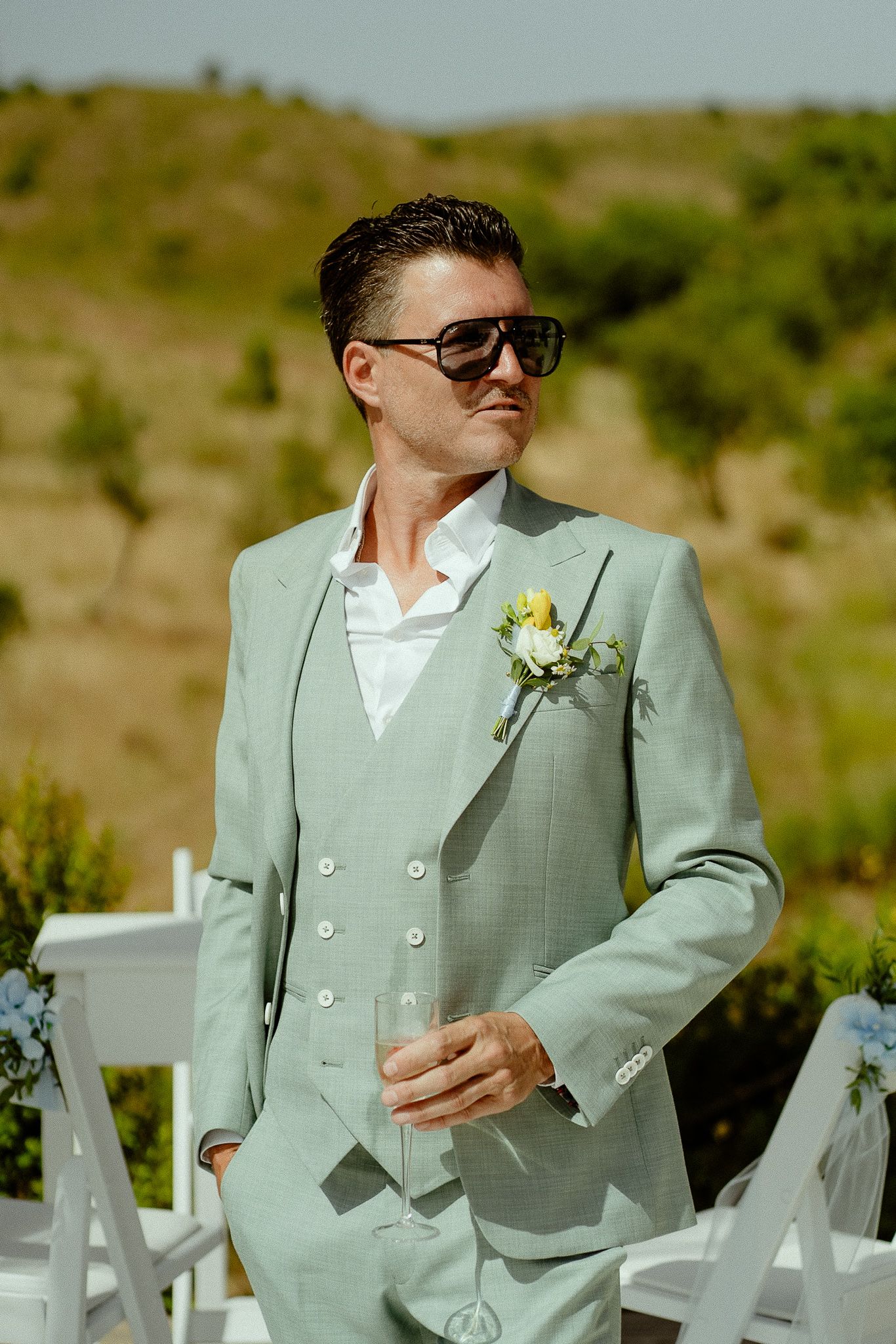 Stylish groom in mint green suit with boutonniere and sunglasses standing outdoors at wedding ceremony.
