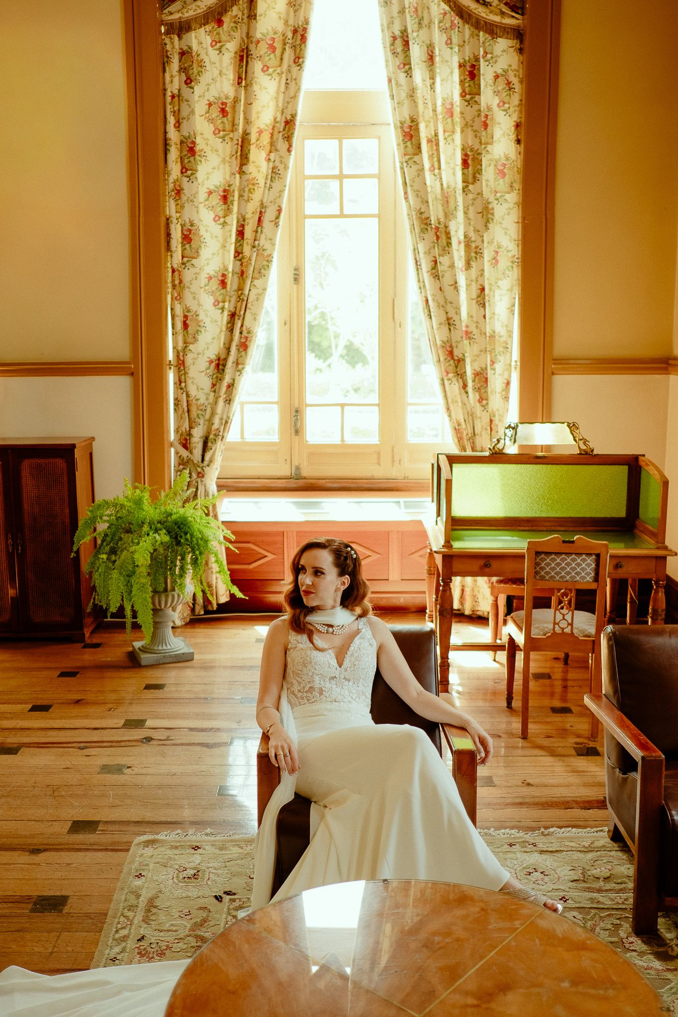 Bride in white lace gown seated in vintage chair inside elegant room with floral curtains and hardwood floors.