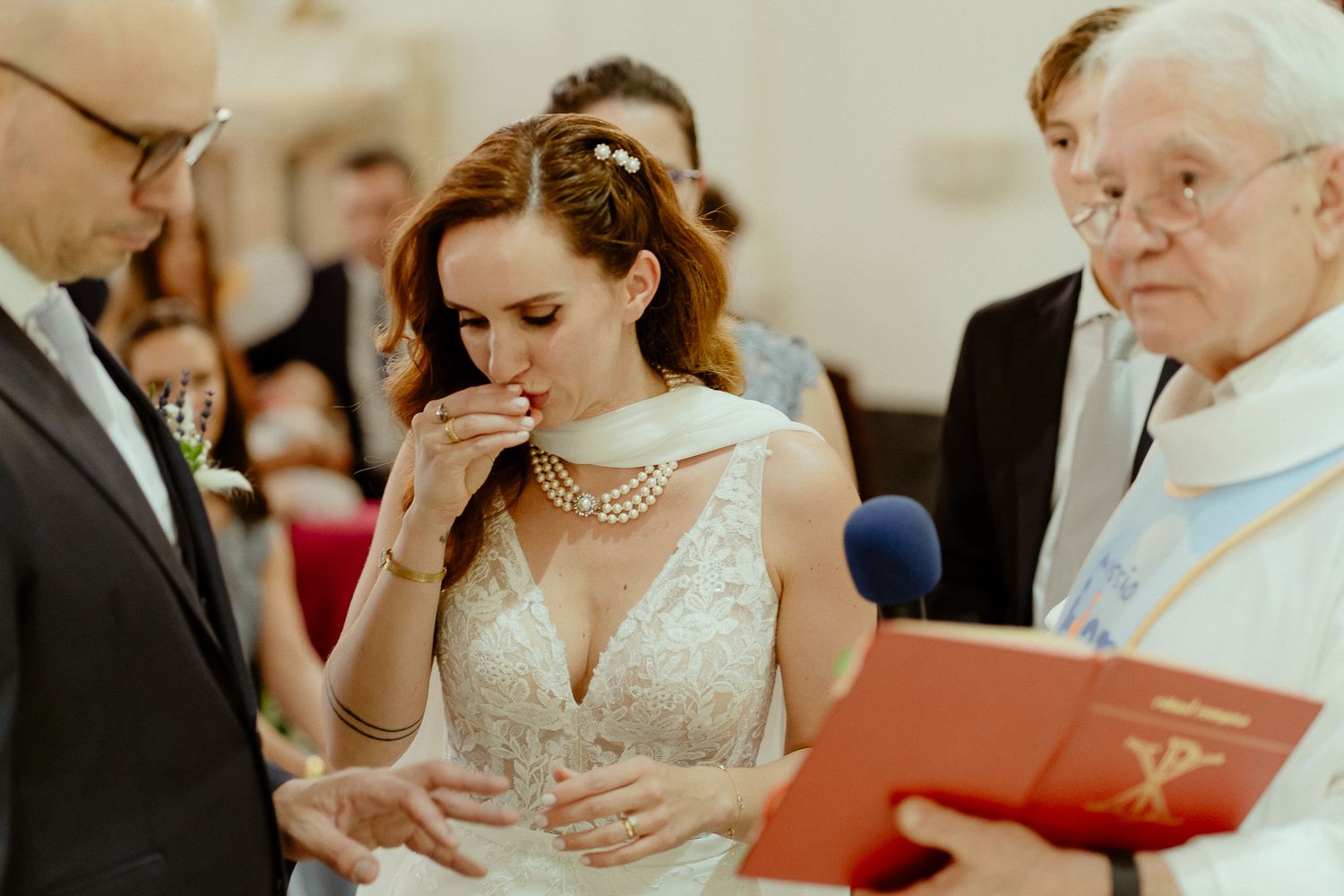Emotional bride in lace wedding gown wiping tears during church ceremony while officiant holds red book.