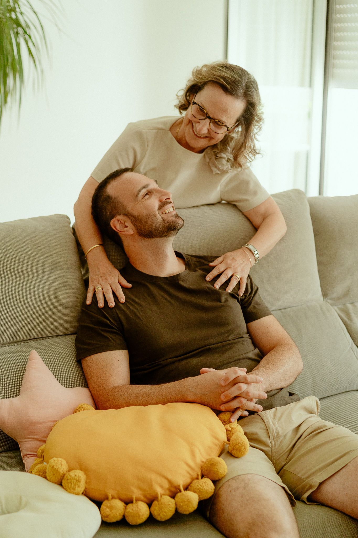 Happy couple relaxing on a cozy sofa, woman with glasses smiling at man leaning back on yellow pillow.