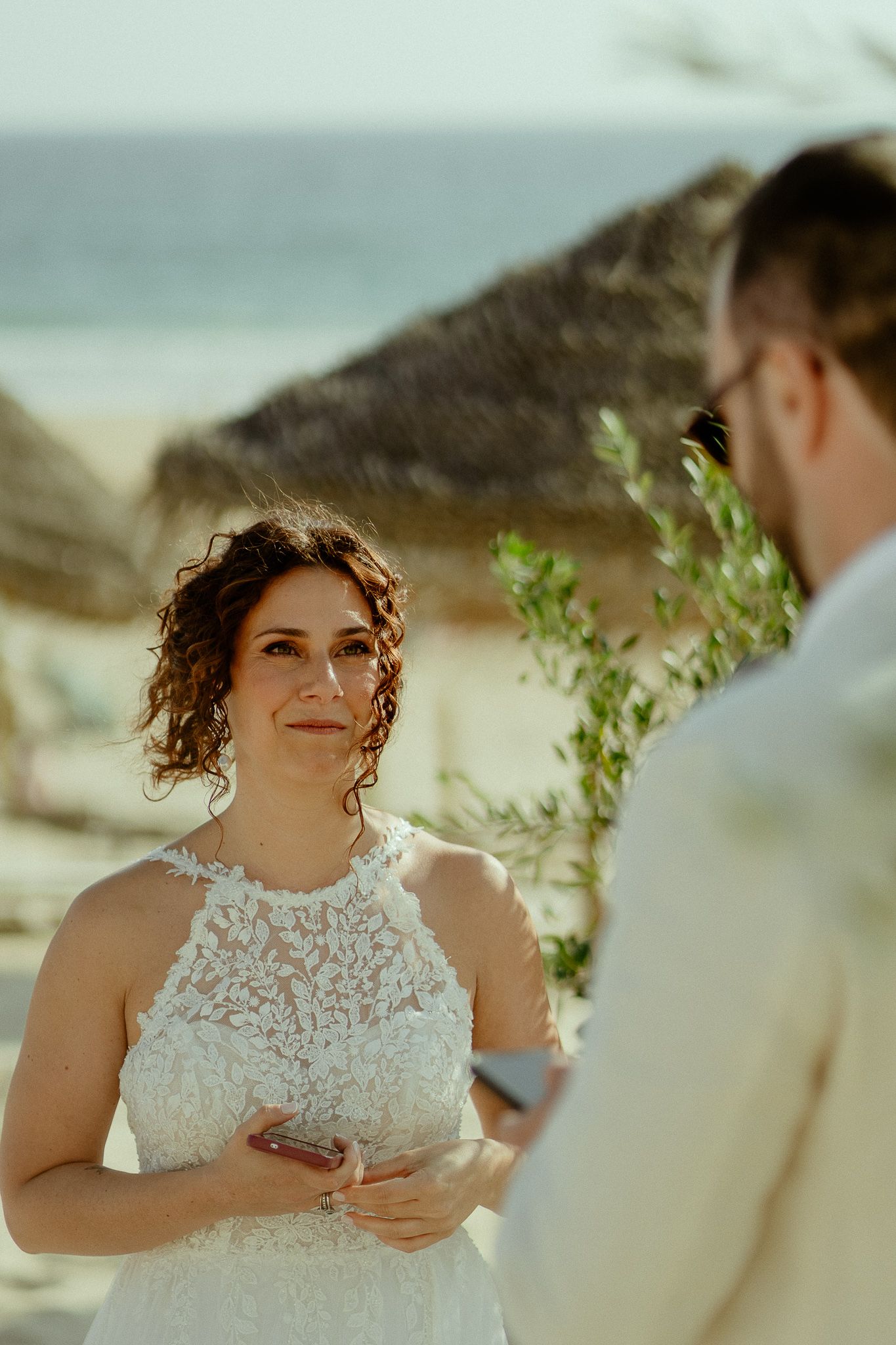 Bride in white lace dress reading vows to groom during romantic beach wedding ceremony.