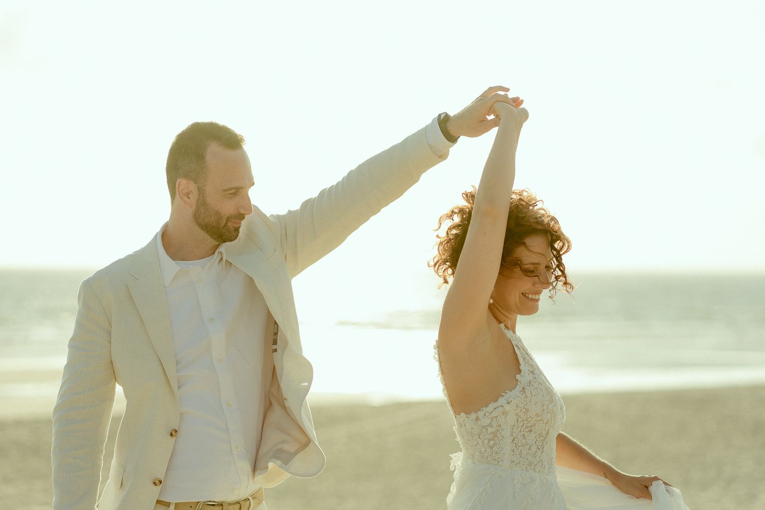 Couple dancing on beach in wedding attire, groom spinning bride in white dress, sunny coastal backdrop.