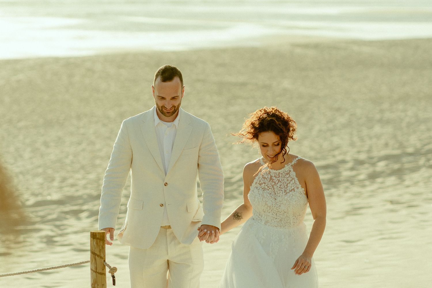Bride and groom hold hands walking on sandy beach at golden sunset in elegant wedding attire.