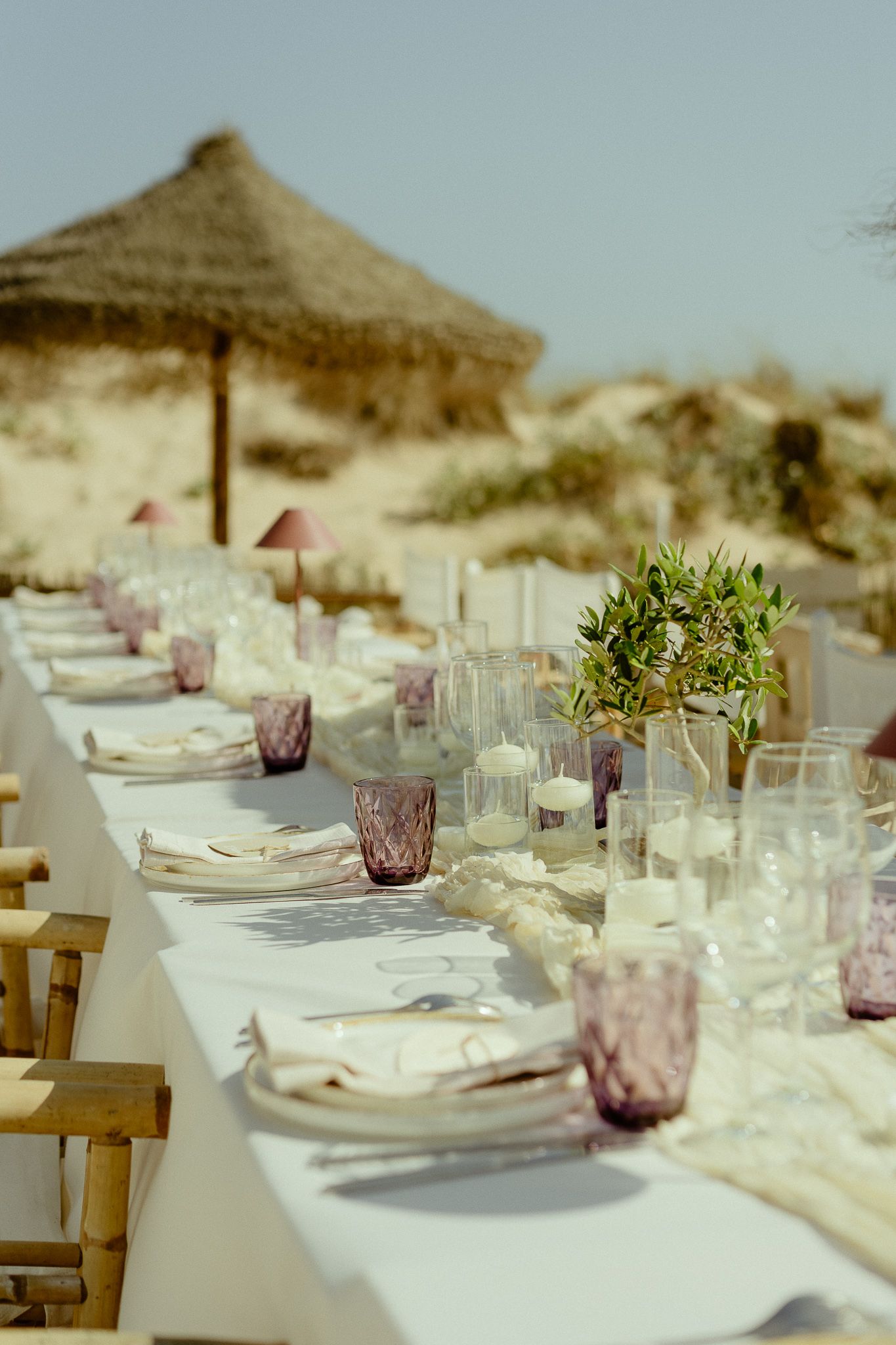 Elegant beach dining table set with purple glasses, candles, and greenery centerpieces under thatched umbrellas.