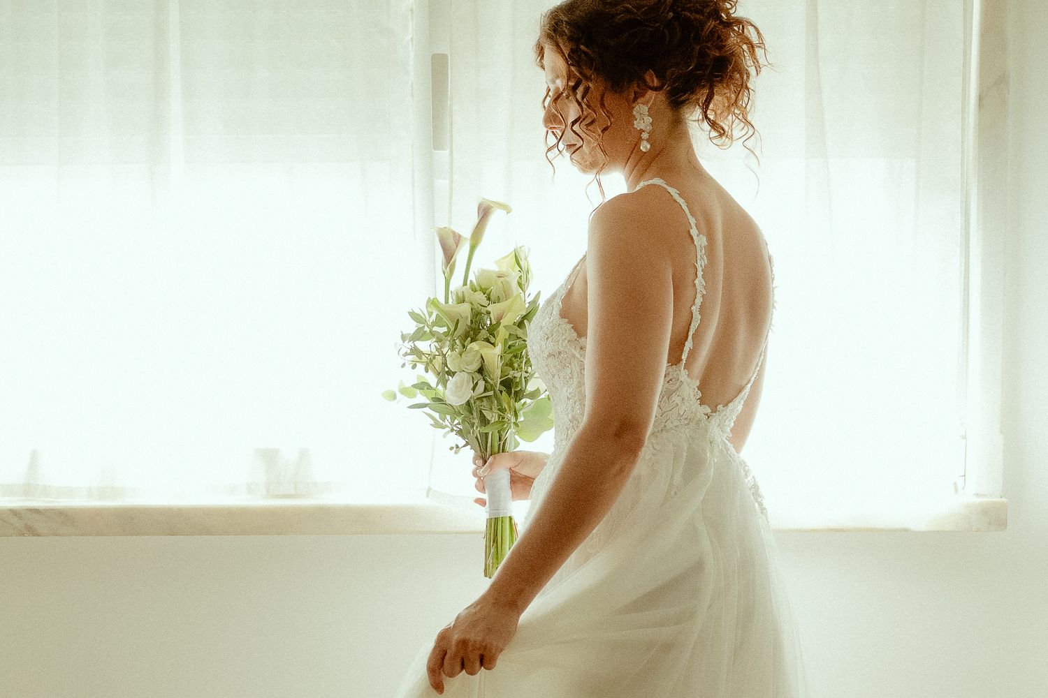 Bride in a white backless wedding gown holding a bouquet of white flowers, standing by a bright window.