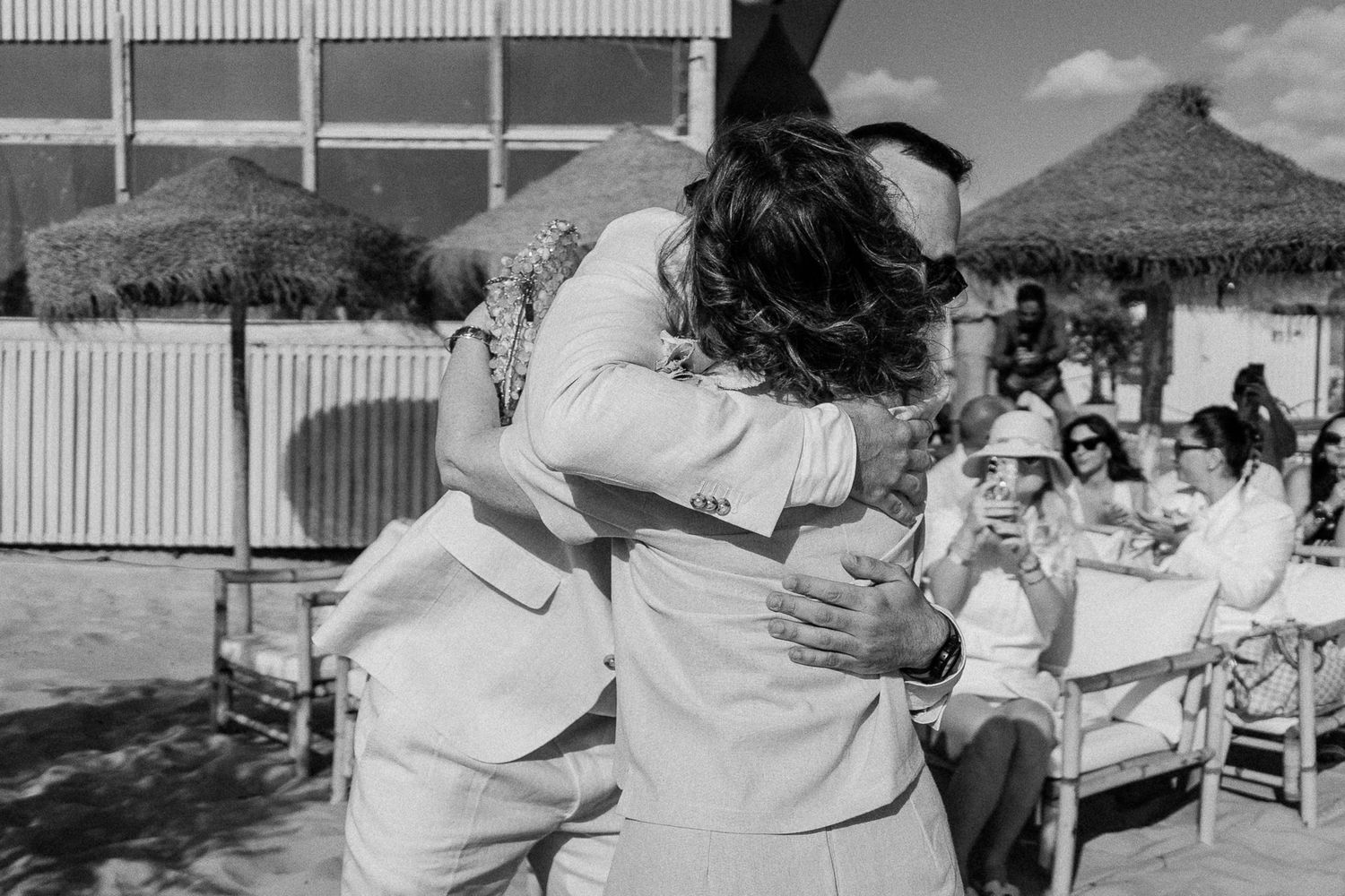 Two people share a heartfelt hug at an outdoor beach wedding ceremony in black and white photo.