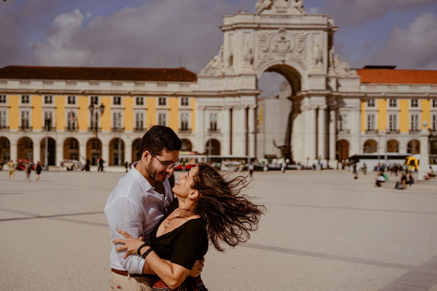 A couple embraces in a passionate kiss at Commerce Square in Lisbon, Portugal, with historic buildings in the background.