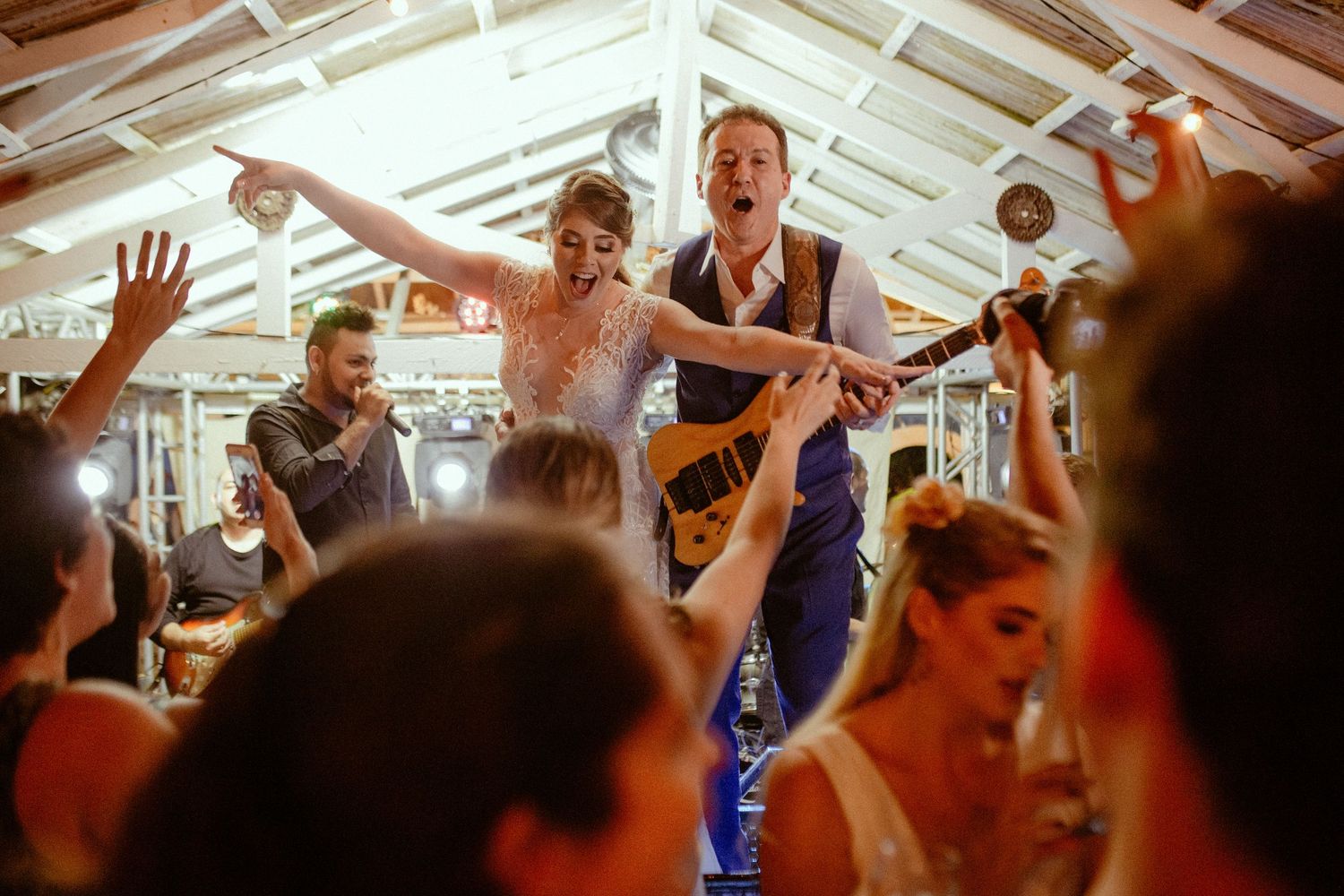 Musicians perform energetically on stage at a crowded indoor venue as audience members raise their hands in celebration.