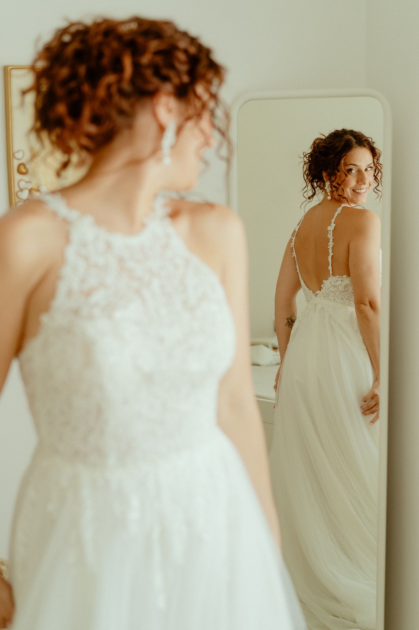 Bride in white lace halter gown smiles at her reflection in a full-length mirror on her wedding day.