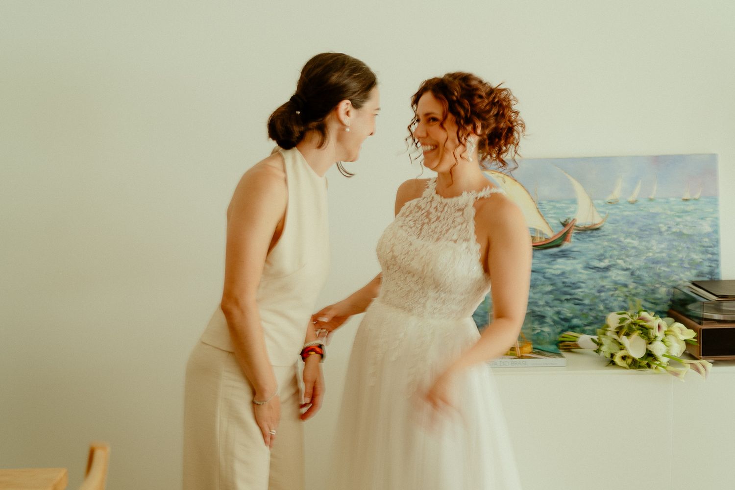 Bride in lace gown and woman in white outfit share a joyful moment near nautical painting.