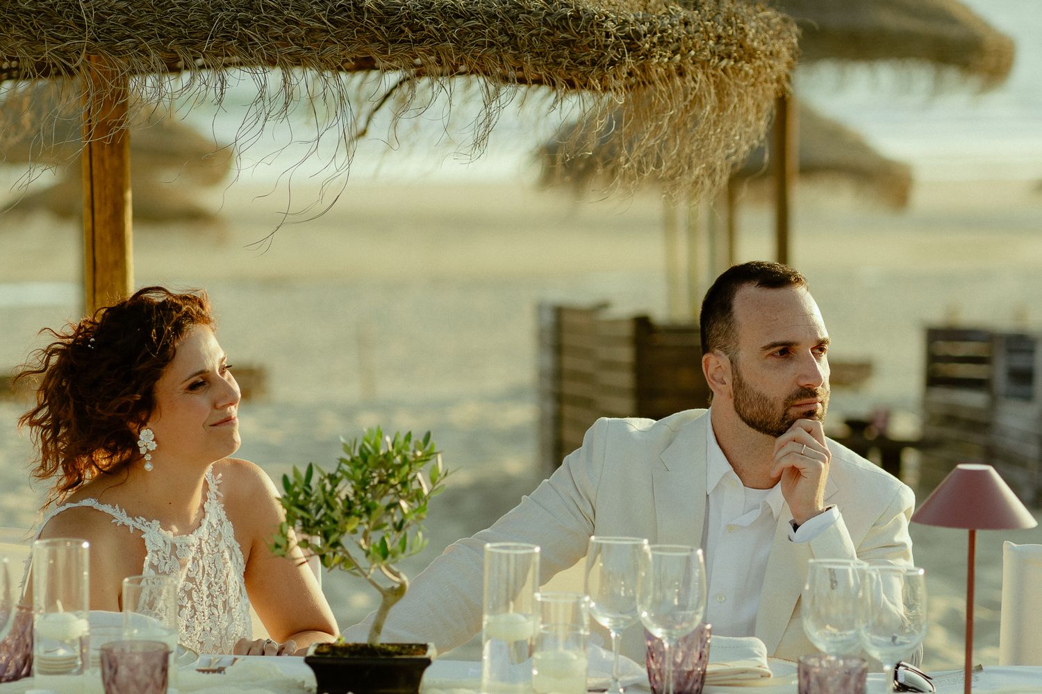 Couple in elegant attire seated at a beachside dining table under thatched umbrellas during an outdoor event.