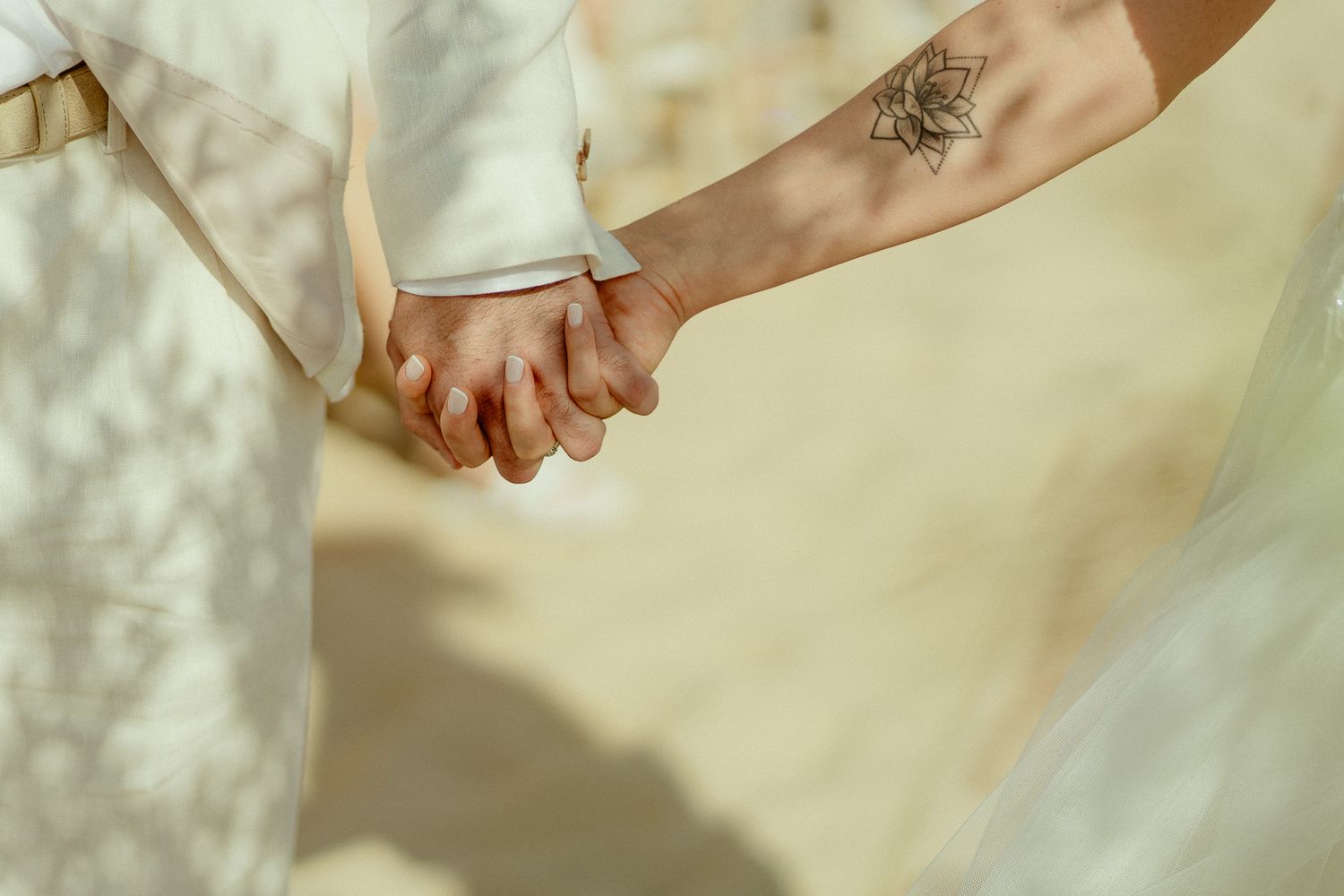 Close-up of a couple holding hands, one with a floral tattoo, wearing wedding attire in soft natural light.