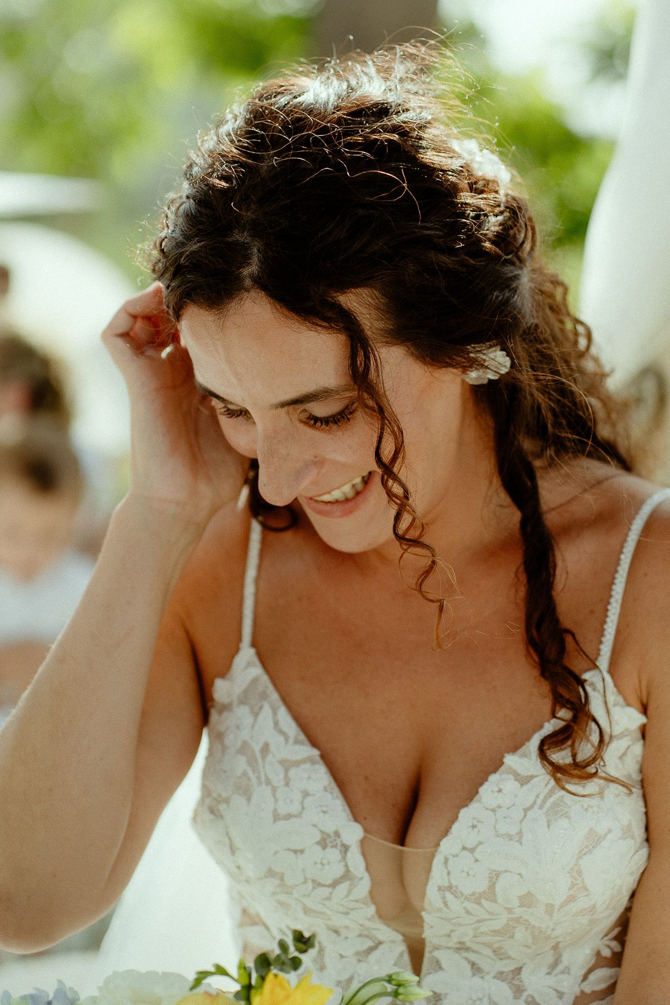 Smiling bride in lace wedding dress with curly hair adorned with flowers, looking down joyfully.