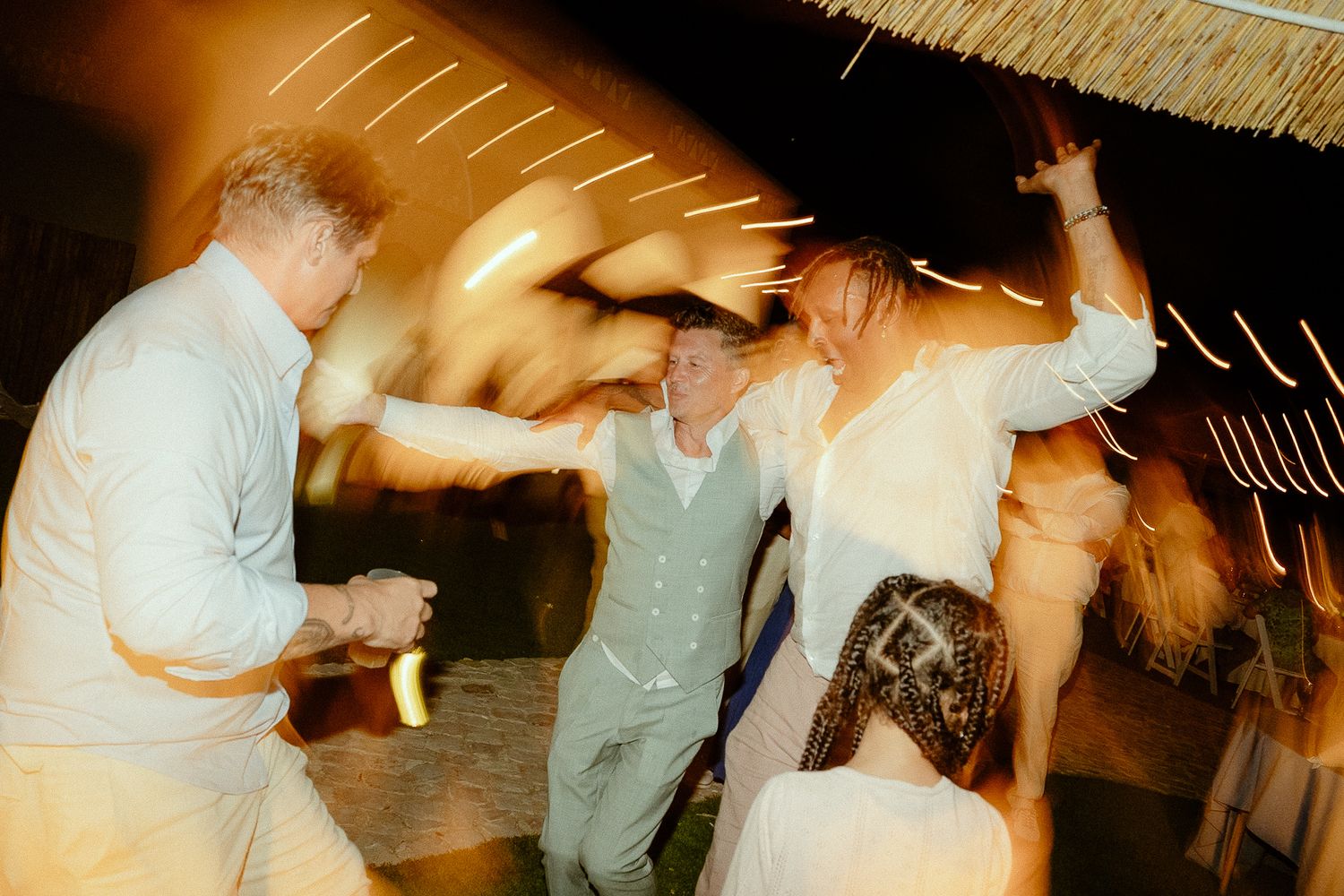Guests dancing energetically at a wedding reception party under thatched roof with motion blur effect.