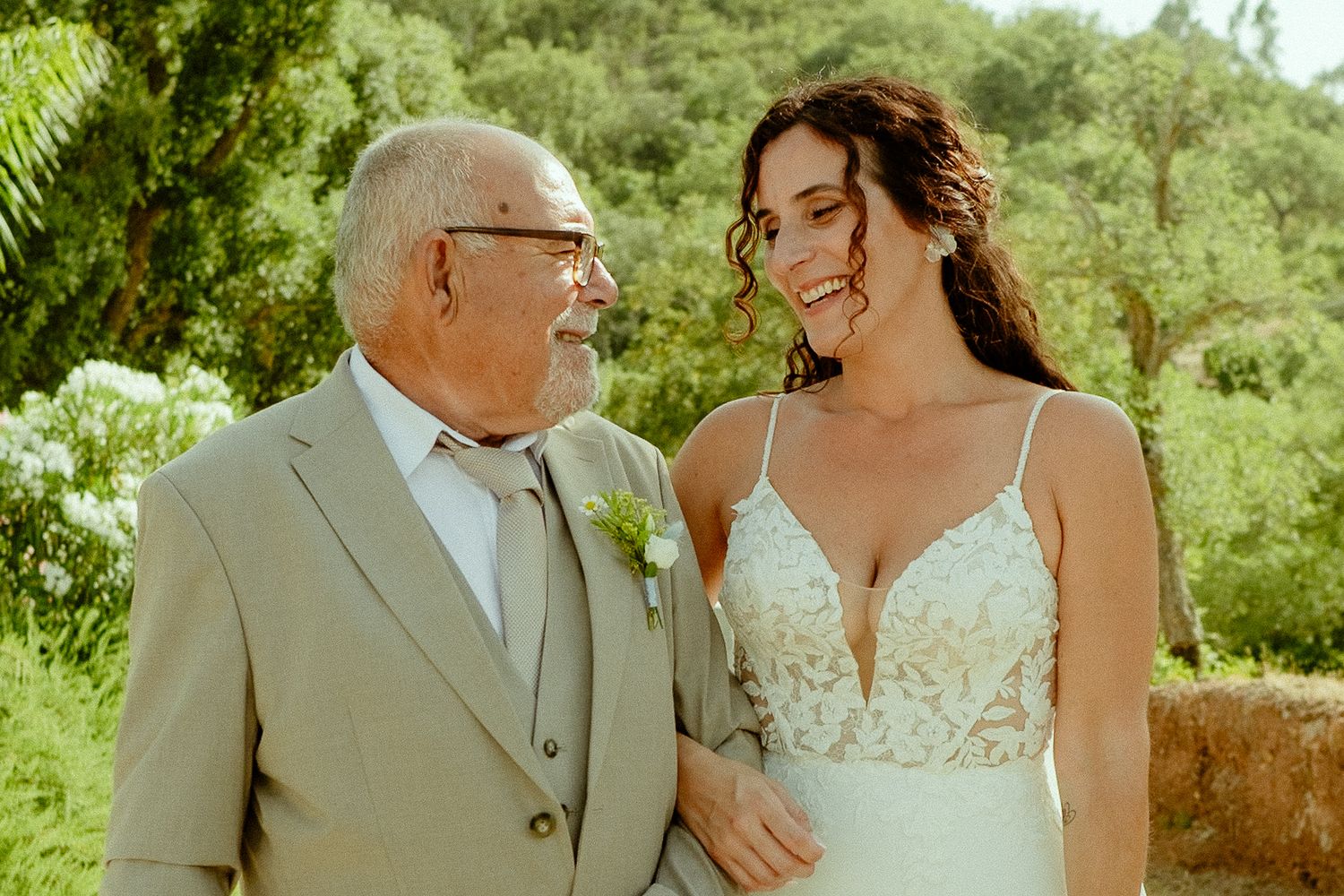 Elderly man in gray suit and bride in white lace wedding gown smiling at each other outdoors.