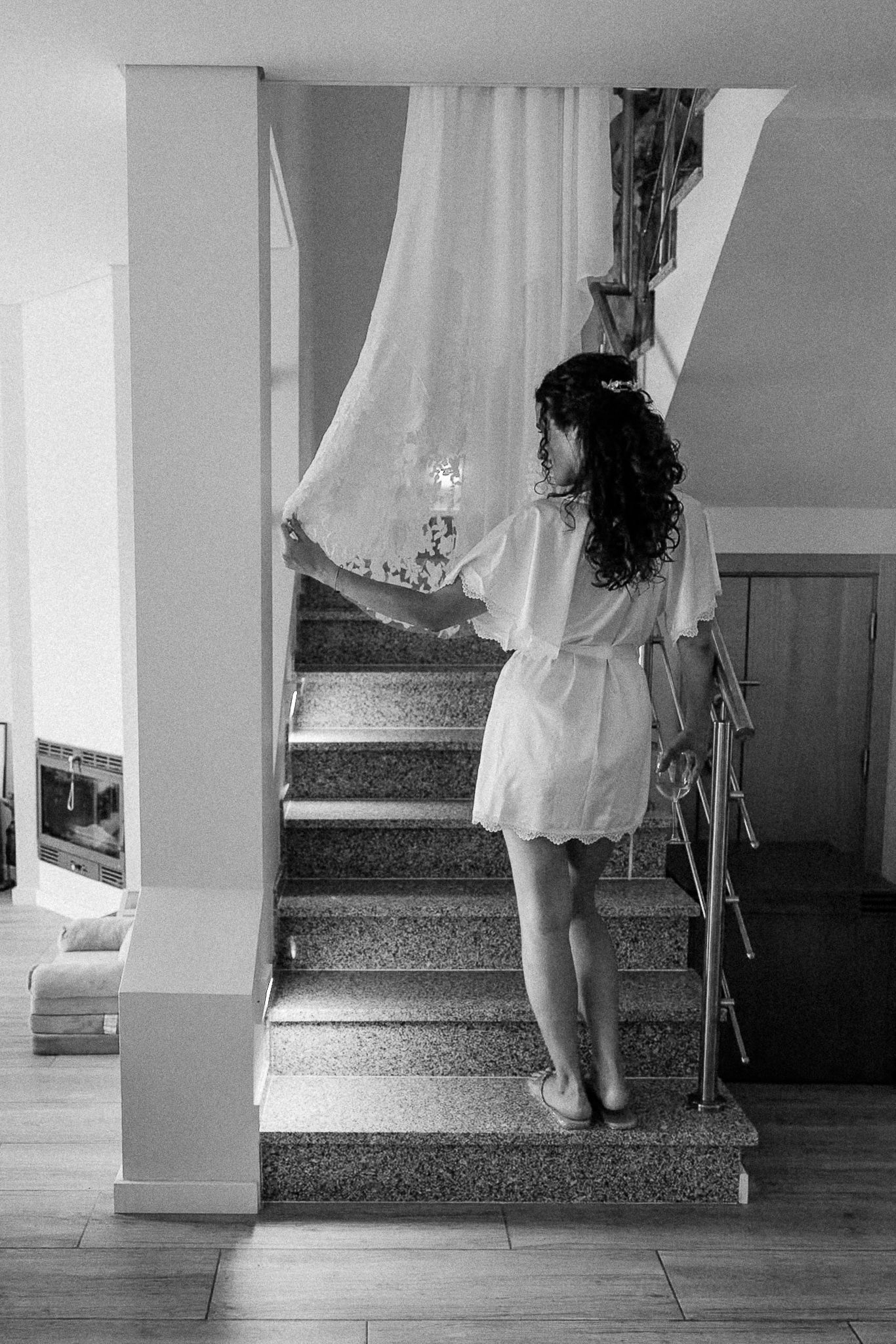 Black and white photo of a woman in a white robe ascending marble stairs, holding a sheer curtain.
