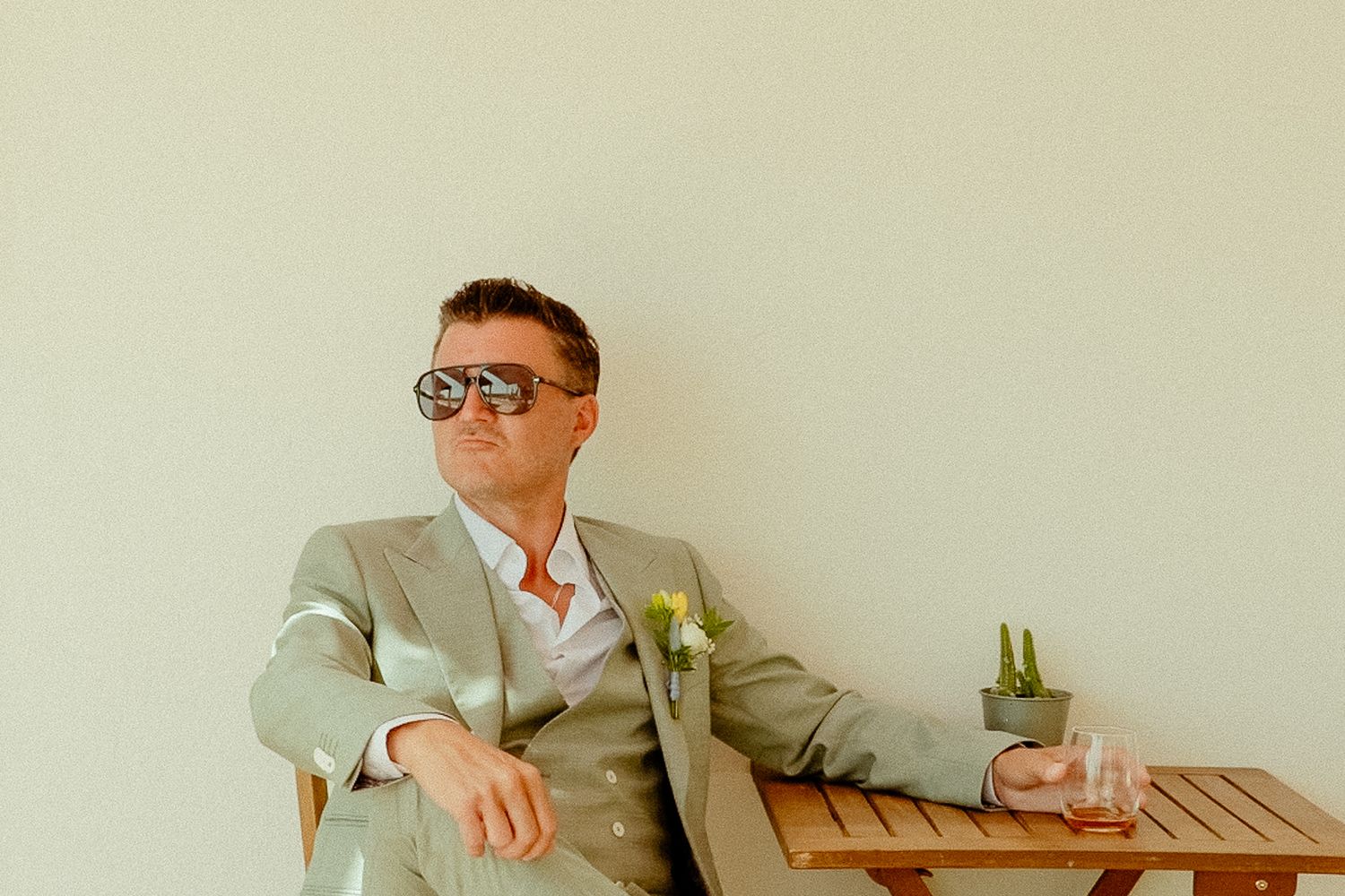 Man in light gray suit and sunglasses relaxing at wooden table holding a glass outdoors.