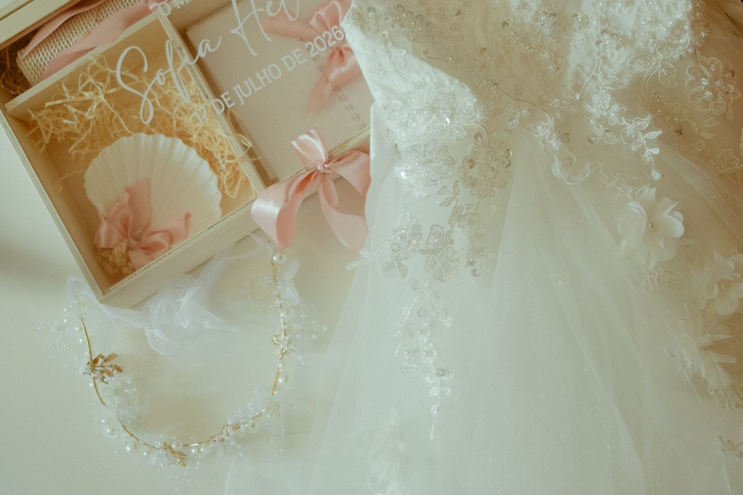 Elegant wedding dress with lace details beside a wooden box with pink ribbon gifts and a delicate gold bridal hair accessory.