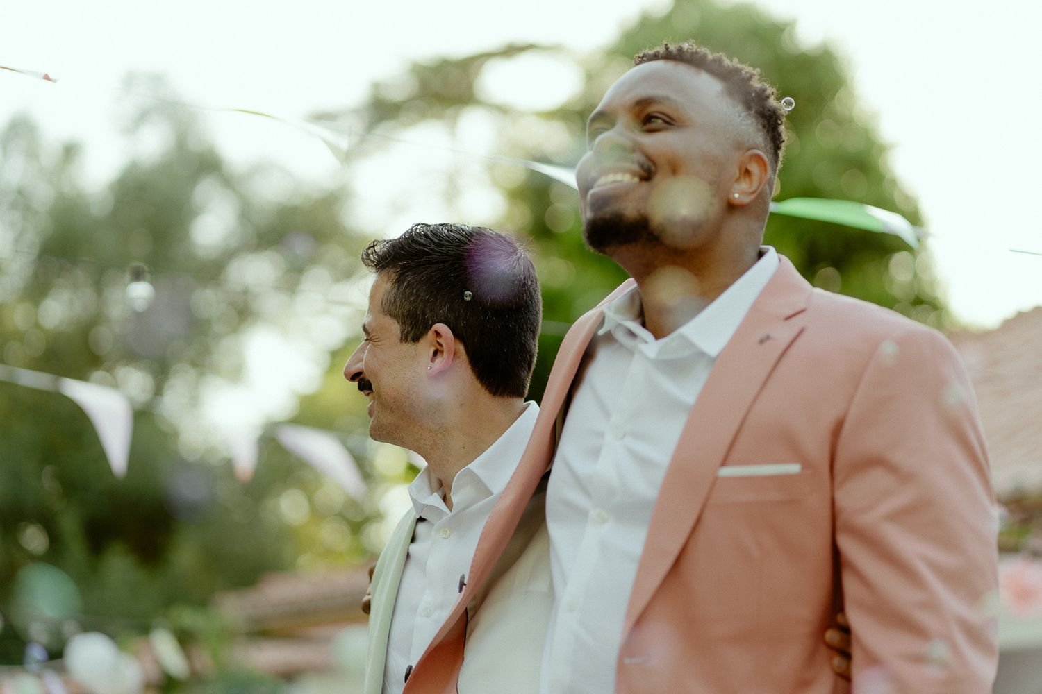 Two men in formal attire, one in a pink blazer, share a tender moment outdoors at what appears to be a wedding.