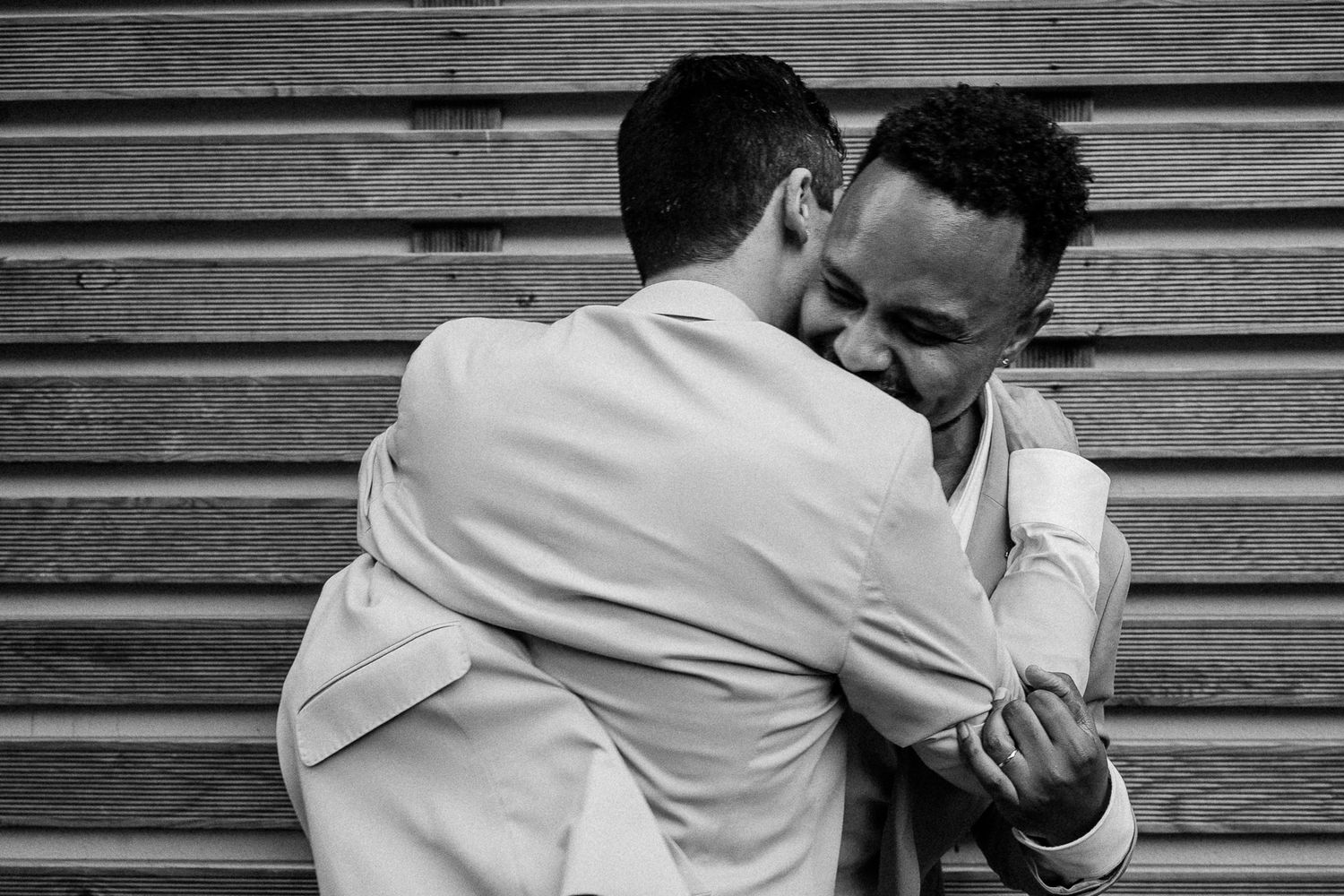 Two grooms in white suits share an emotional embrace on their wedding day in a black and white photo.