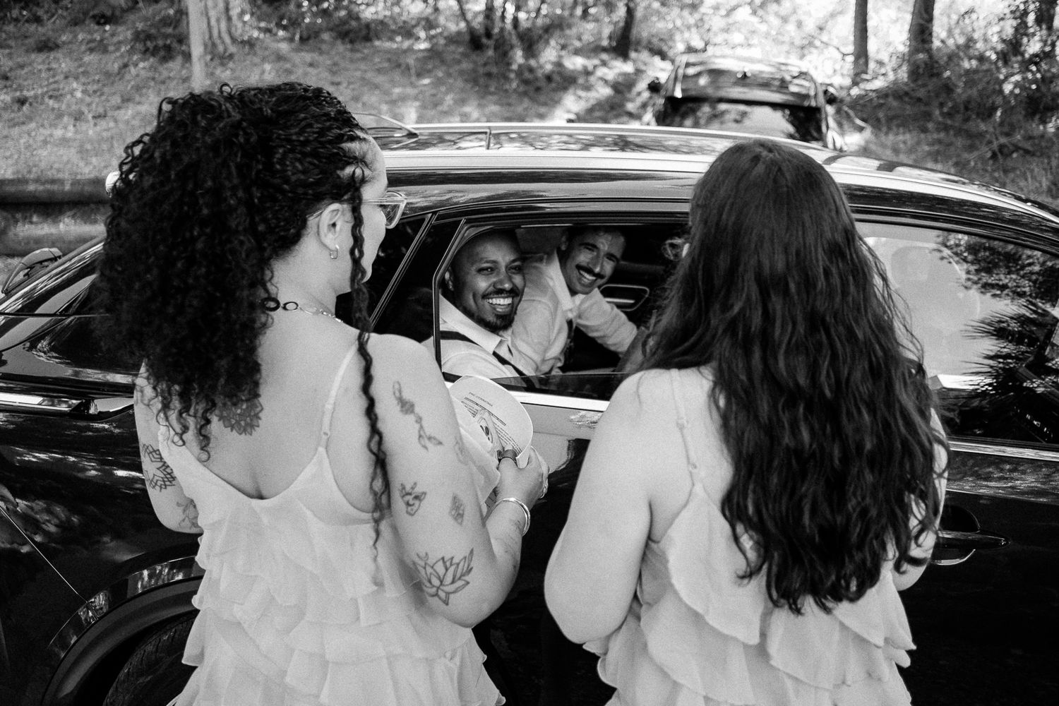 Black and white photo of a man smiling in a car window, flanked by two women in white dresses outdoors.