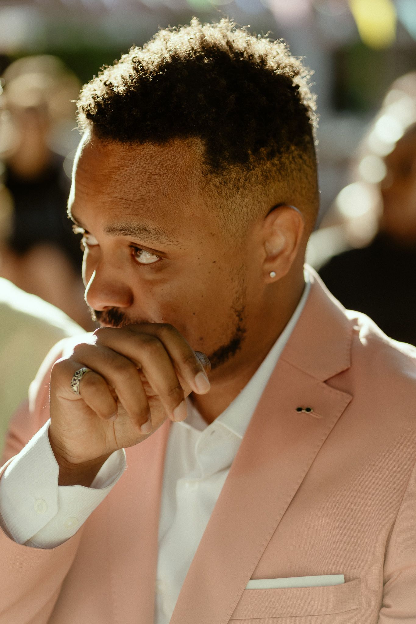A man in a pink blazer and white shirt thoughtfully rests his hand near his mouth outdoors.