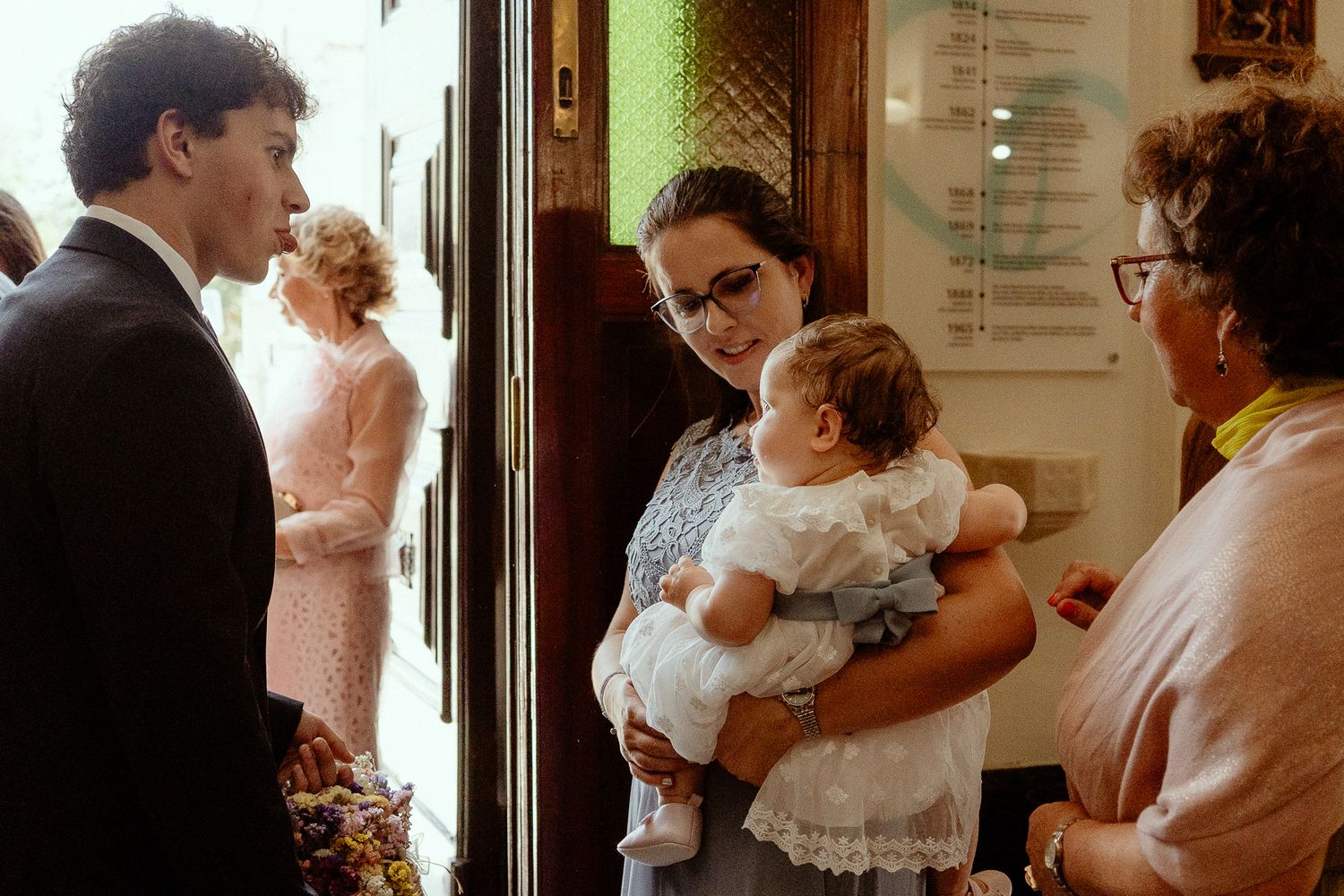 A woman holds a baby in a white christening gown while conversing with guests at a church baptism ceremony.