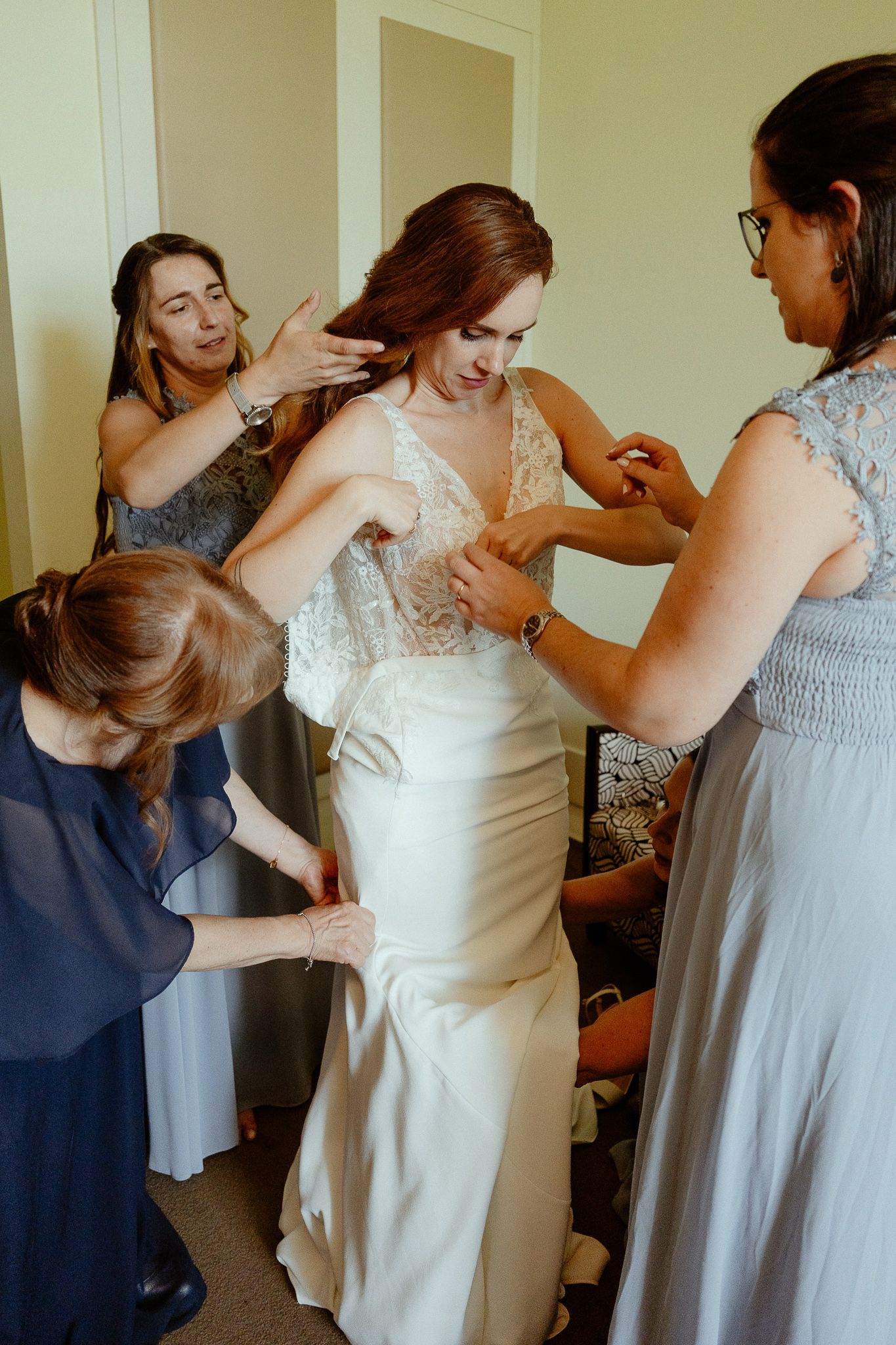 Bridesmaids helping bride get dressed in elegant white lace wedding gown before the ceremony.