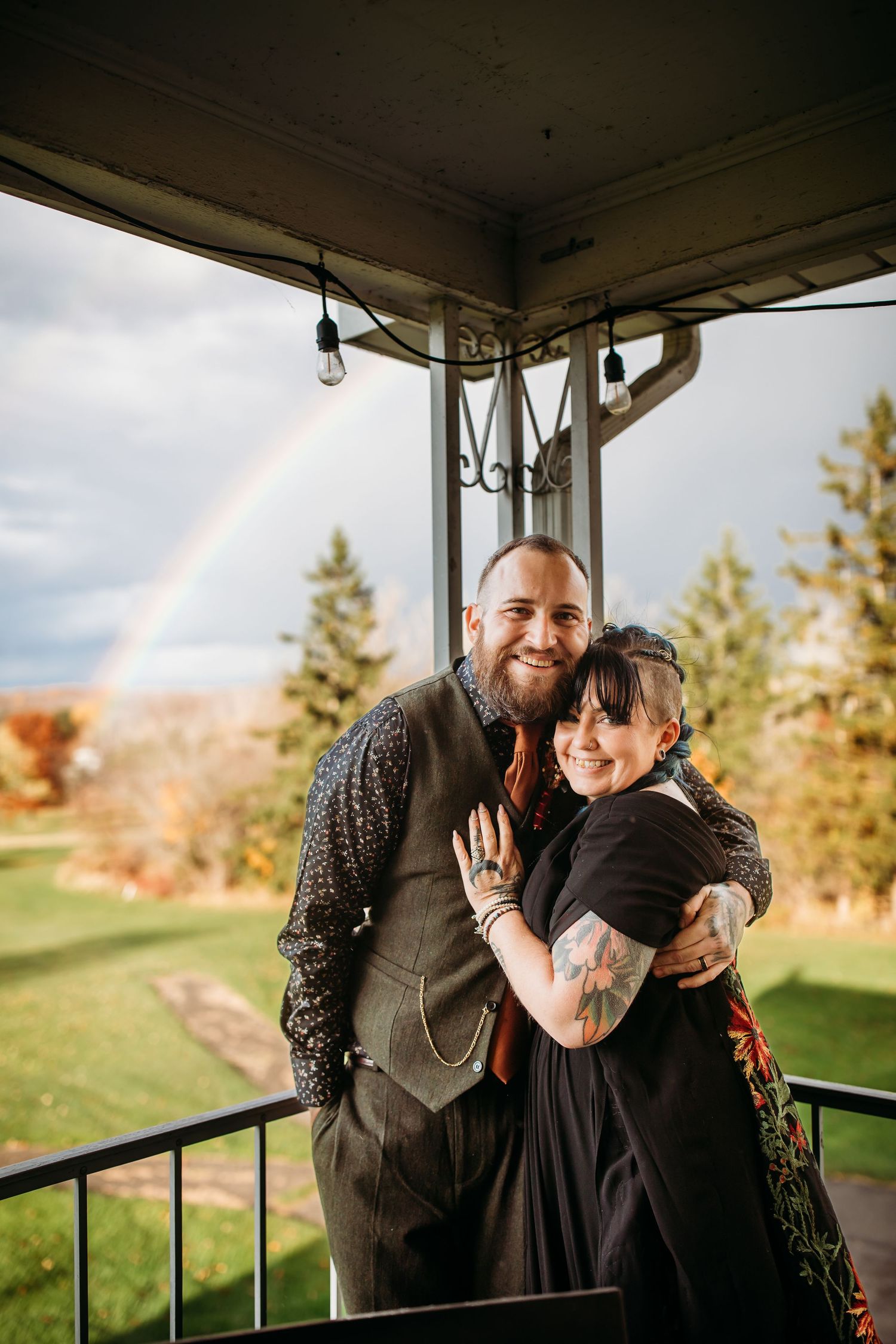 A queer couple embrace in front of a well-timed rainbow on their wedding day.